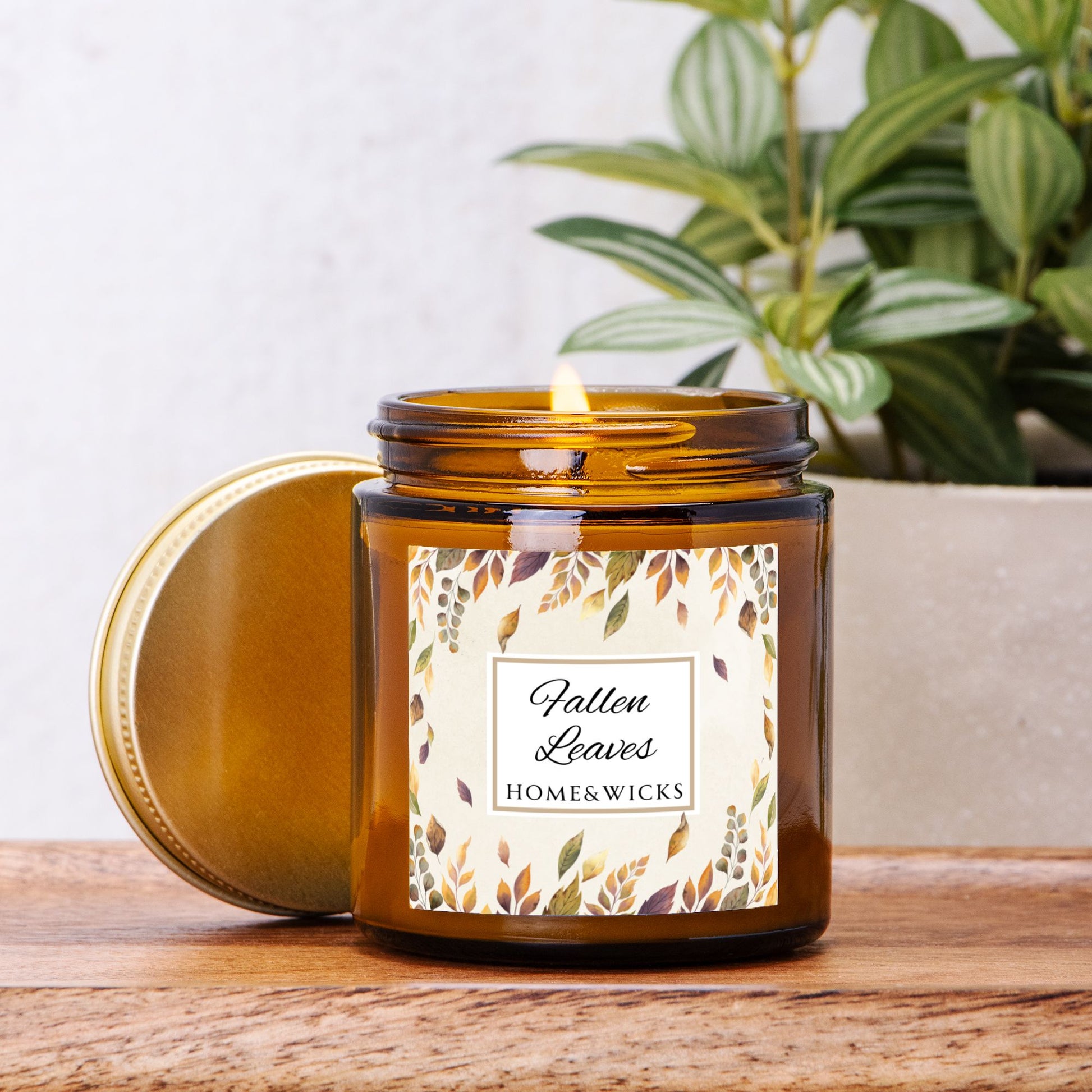 Lit candle labeled 'Fallen Leaves' from Home & Wicks on a wooden surface with a plant in the background.