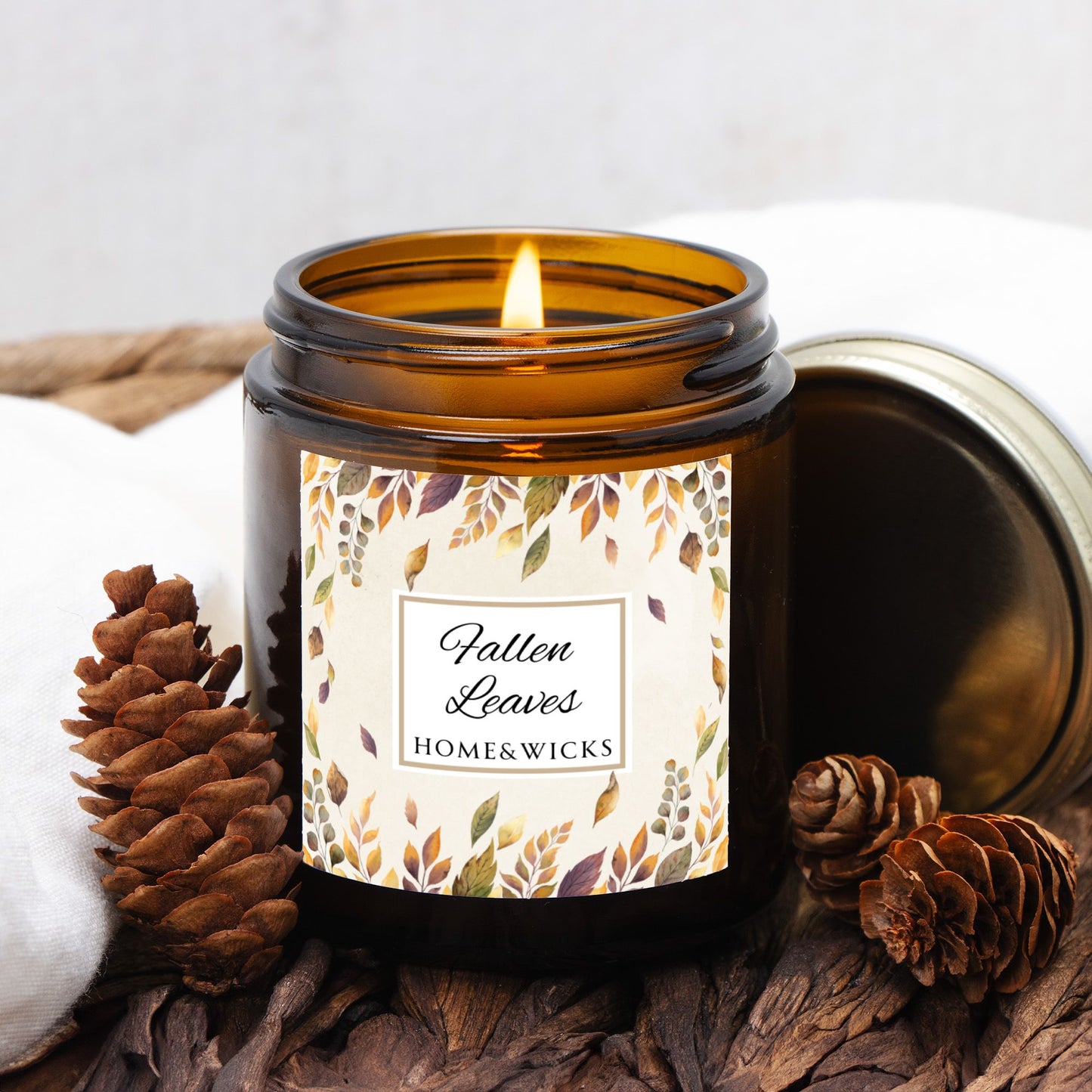 Lit candle in an amber jar labeled 'Fallen Leaves' from Home & Wicks, surrounded by pine cones and wood.