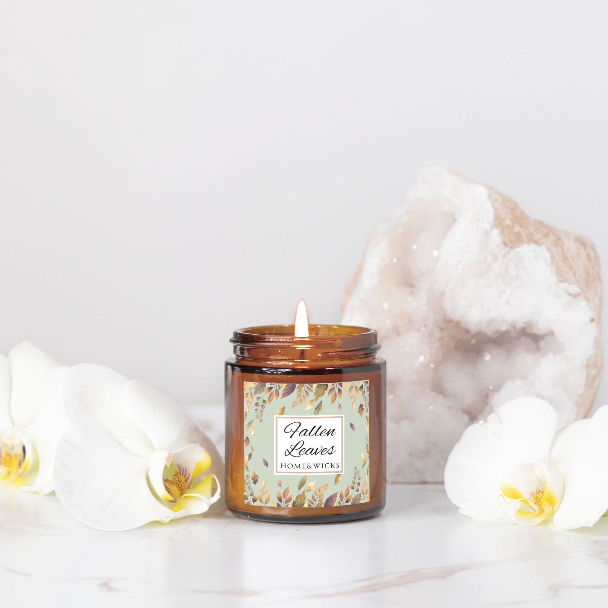 Fallen Leaves candle in a 4 oz. amber glass jar with gold lid with a 'Fallen Leaves' label on a white background by Home & Wicks on a white surface with decorative elements.