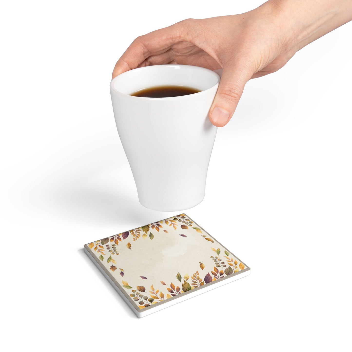 Hand holding a white mug with a Fallen Leaves tile coaster underneath on a white background