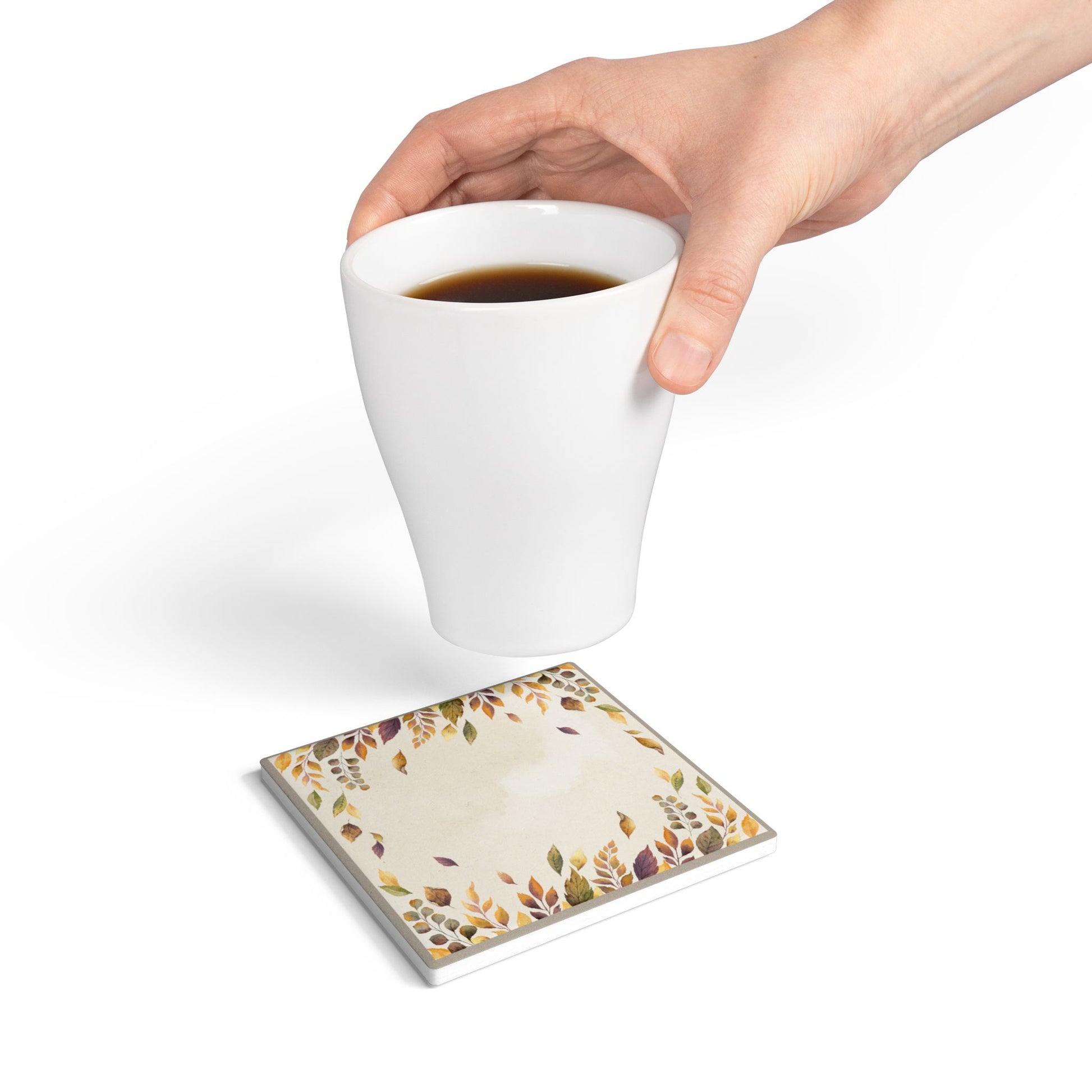 Hand holding a white mug with a Fallen Leaves tile coaster underneath on a white background