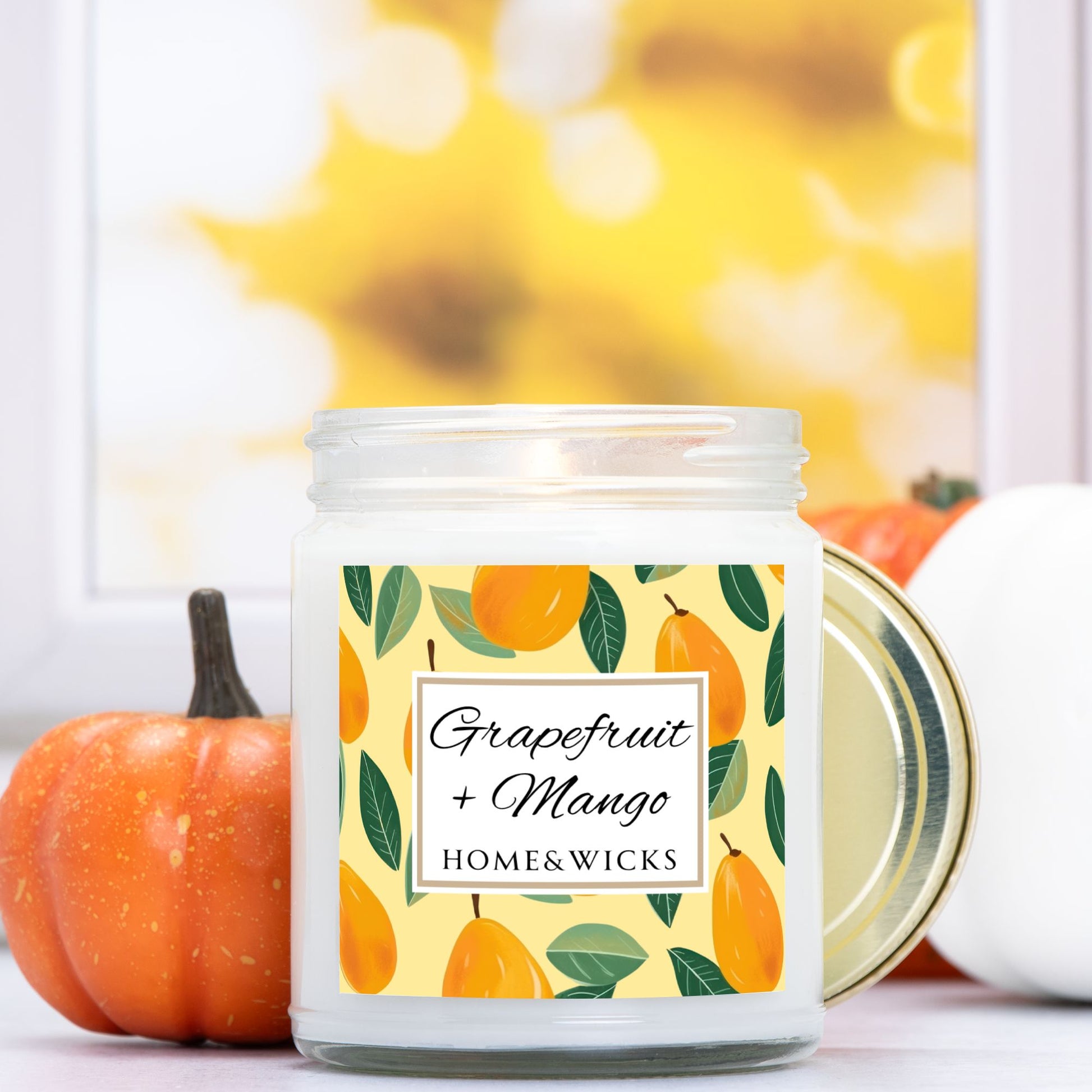 Grapefruit and Mango candle in a 9 oz. clear jar with gold lid and  a decorative mango label on a white background by Home & Wicks on a blurred background with pumpkins
