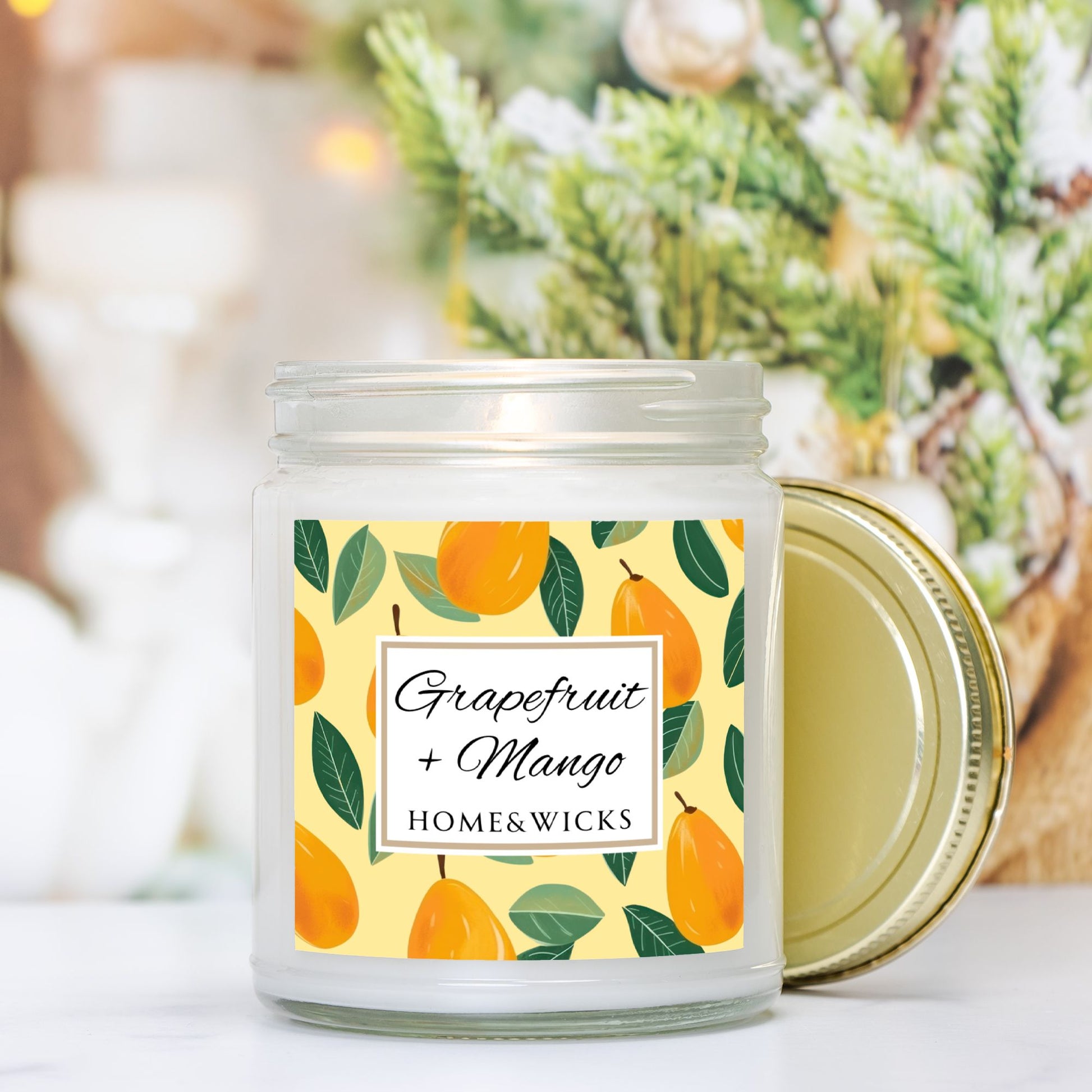 Grapefruit and Mango candle in a 9 oz. clear jar with gold lid and  a decorative mango label on a white background by Home & Wicks on a blurred background with greenery