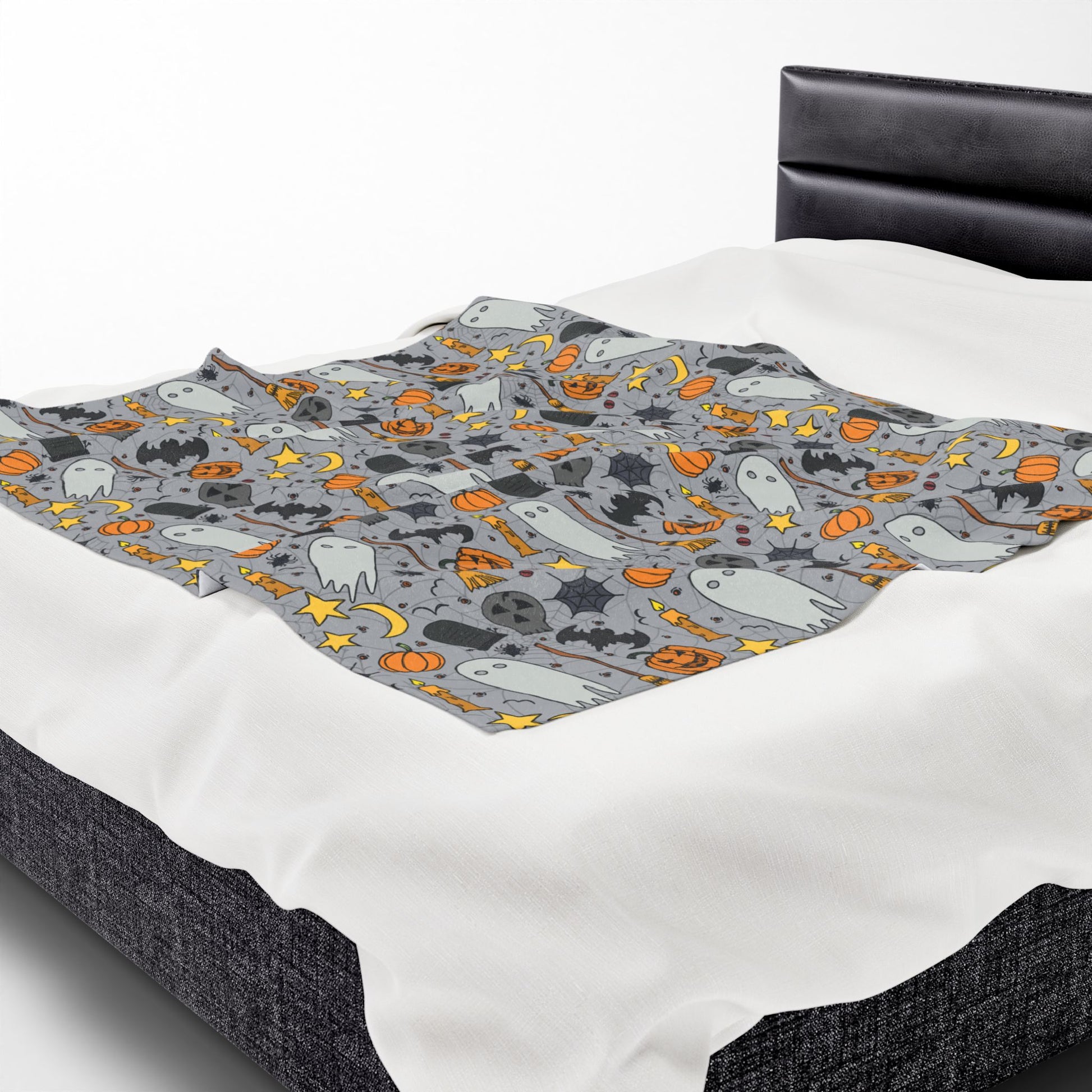 Halloween-themed patterned blanket with ghosts, pumpkins, and stars on a gray background on front of blanket, on a bed.