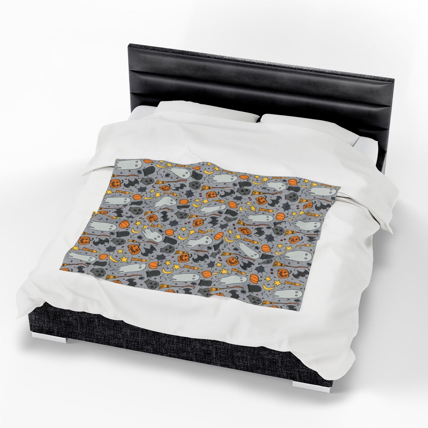 Halloween-themed patterned blanket with ghosts, pumpkins, and stars on a gray background on front of blanket on bed.