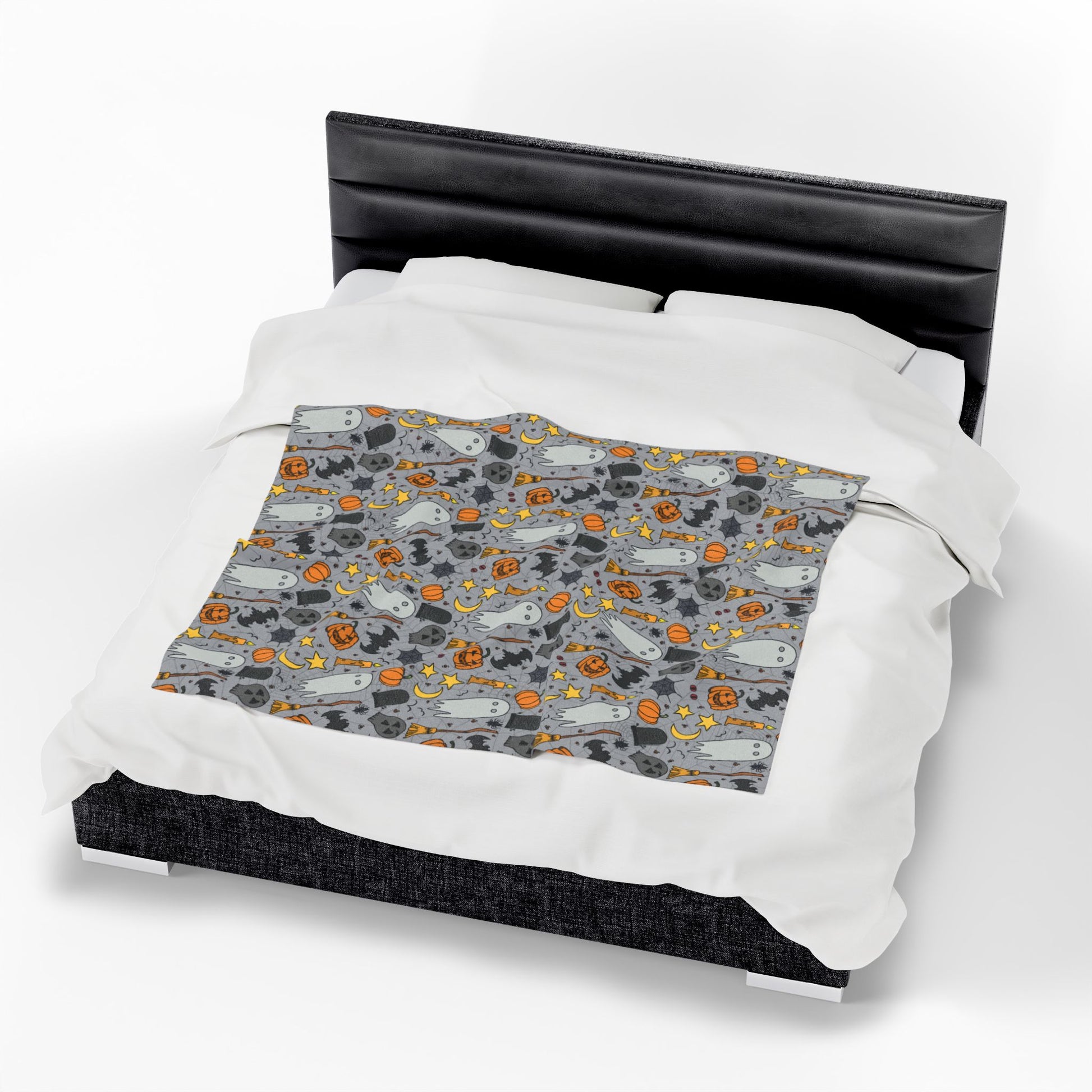 Halloween-themed patterned blanket with ghosts, pumpkins, and stars on a gray background on front of blanket on bed.