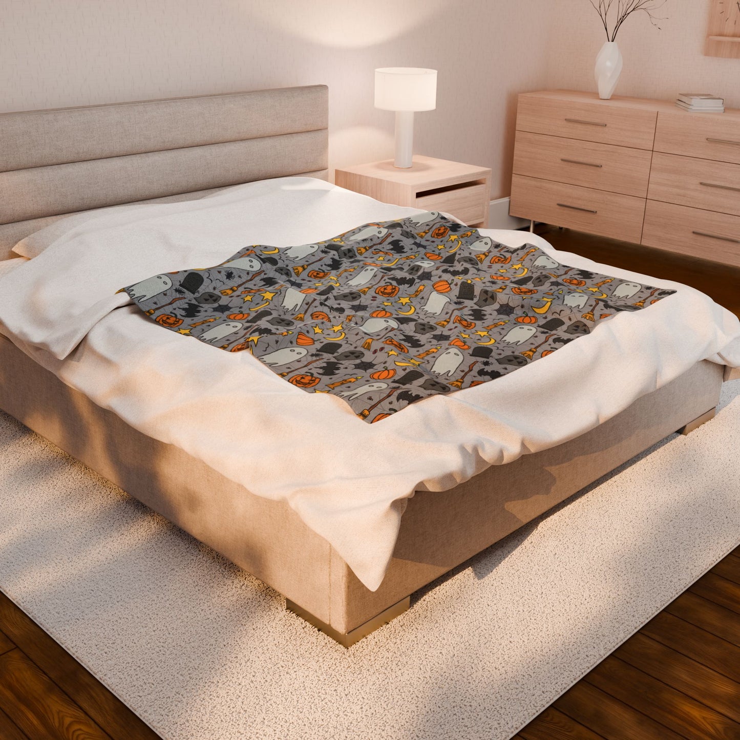 Halloween-themed patterned blanket with ghosts, pumpkins, and stars on a gray background on front of blanket, on bed with a patterned blanket in a bedroom setting.