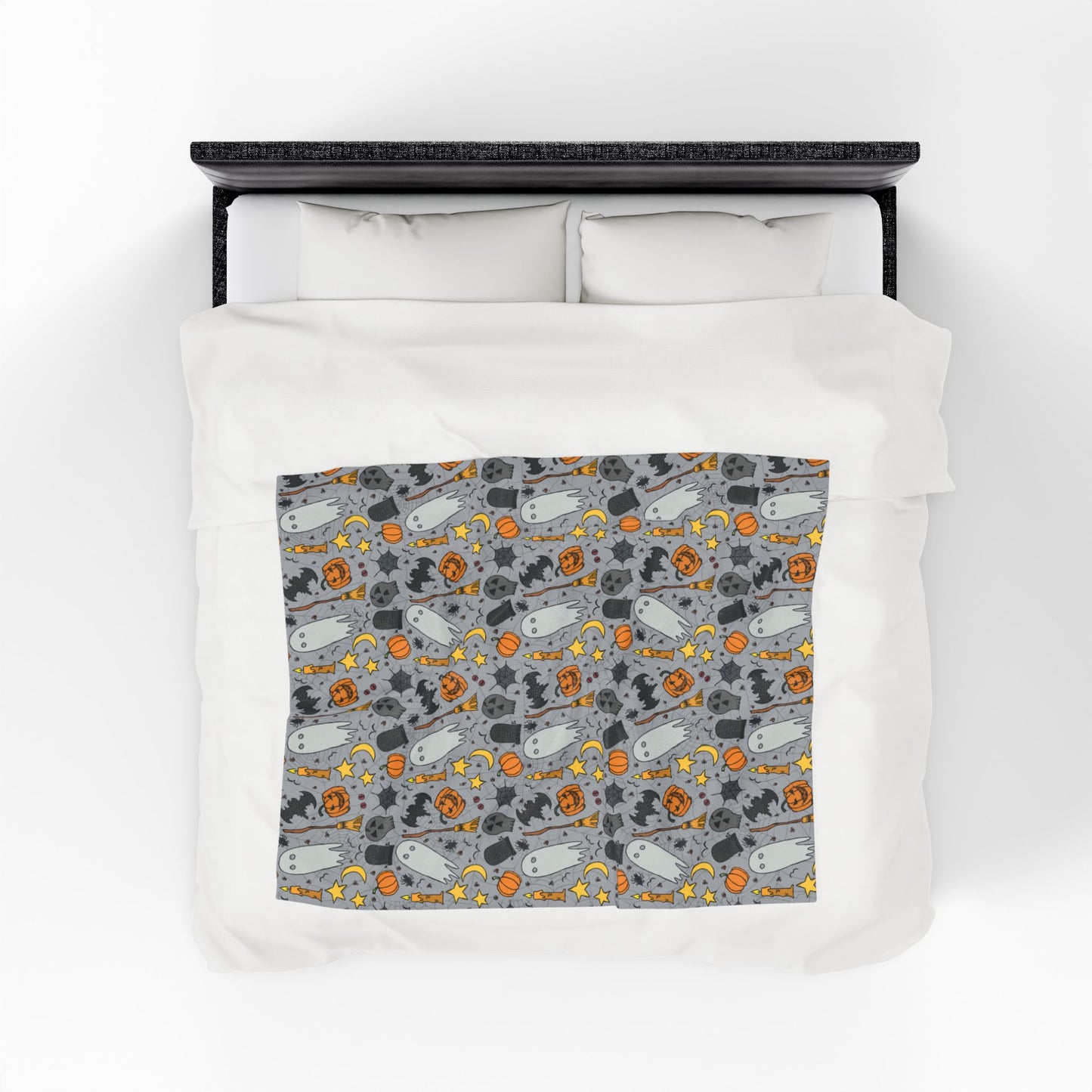 Halloween-themed patterned blanket with ghosts, pumpkins, and stars on a gray background on front of blanket on a bed.
