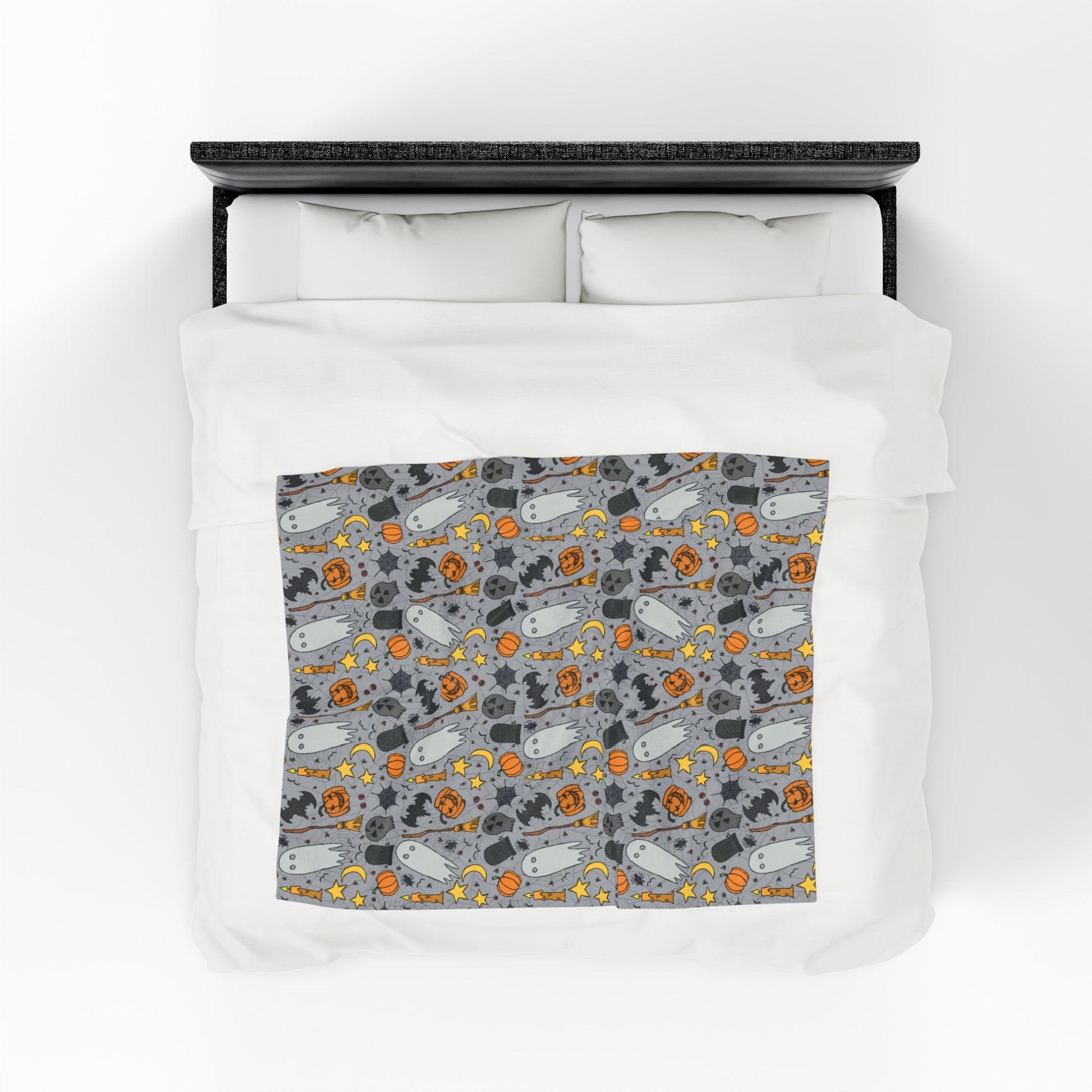 Halloween-themed patterned blanket with ghosts, pumpkins, and stars on a gray background on front of blanket on a bed.