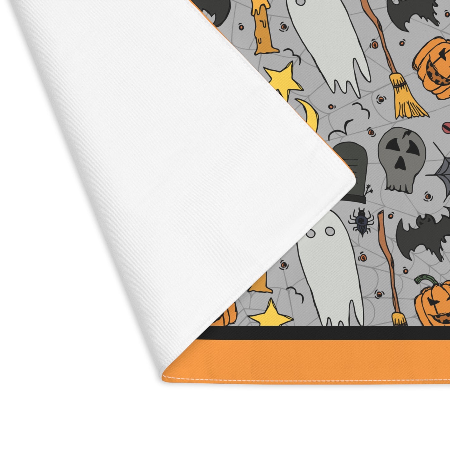 Halloween-themed placemat with ghosts, pumpkins, and bats on a gray background with an orange border, with a corner folded over to reveal white backside of placemat, on a white background.