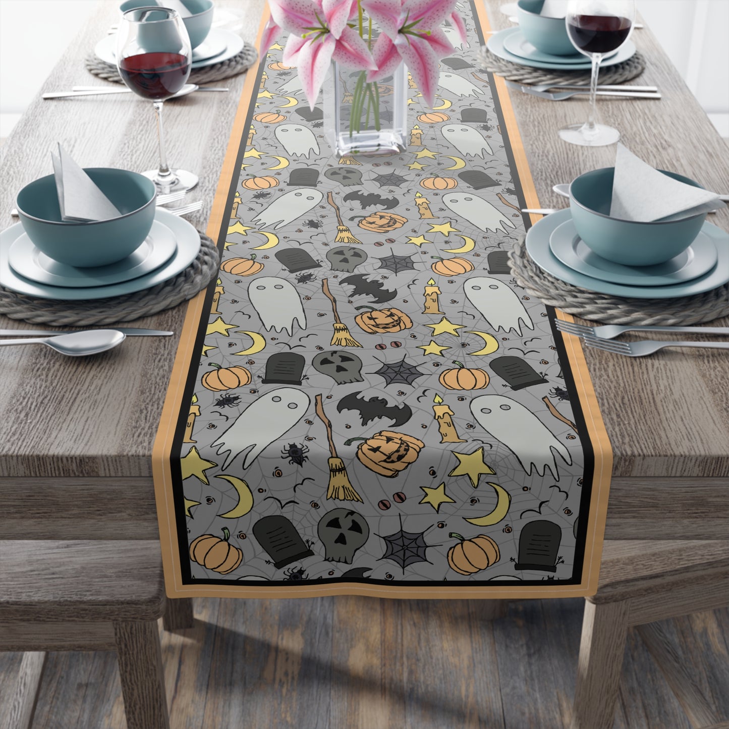 Halloween-themed table runner with ghosts, pumpkins, and bats with a gray background on front of table runner, on a wooden table with dining setup.