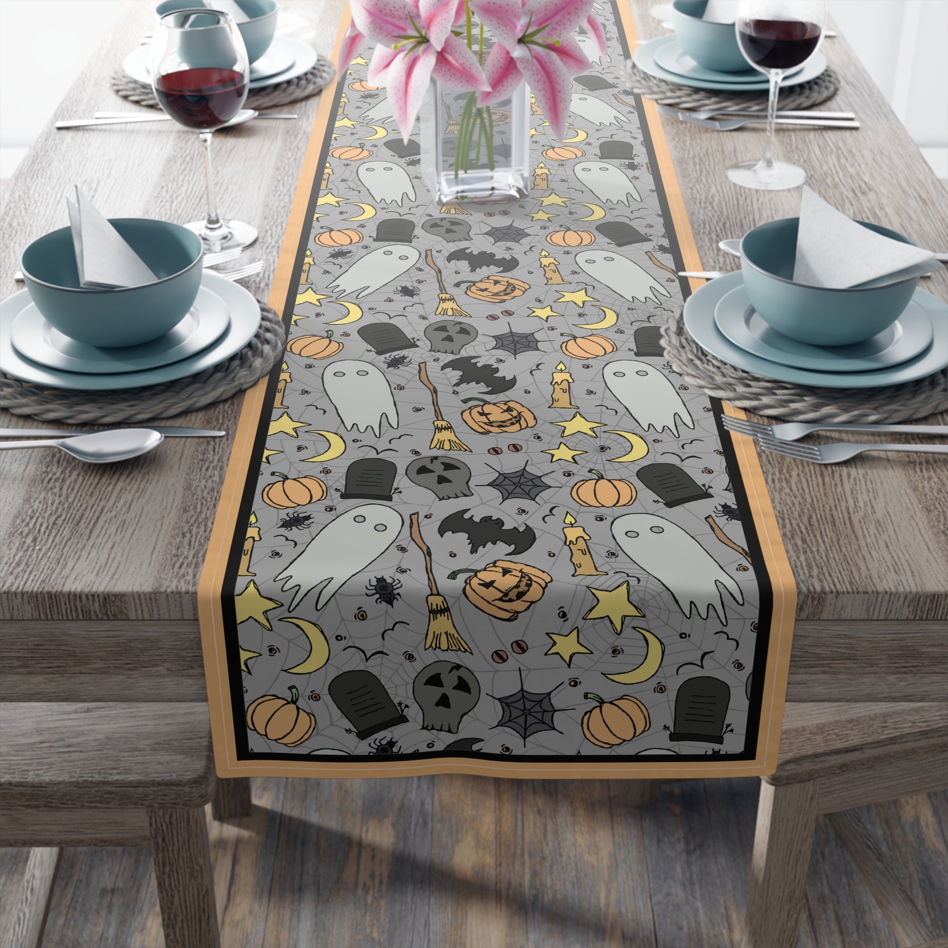 Halloween-themed table runner with ghosts, pumpkins, and bats with a gray background on front of table runner, on a wooden table with dining setup.