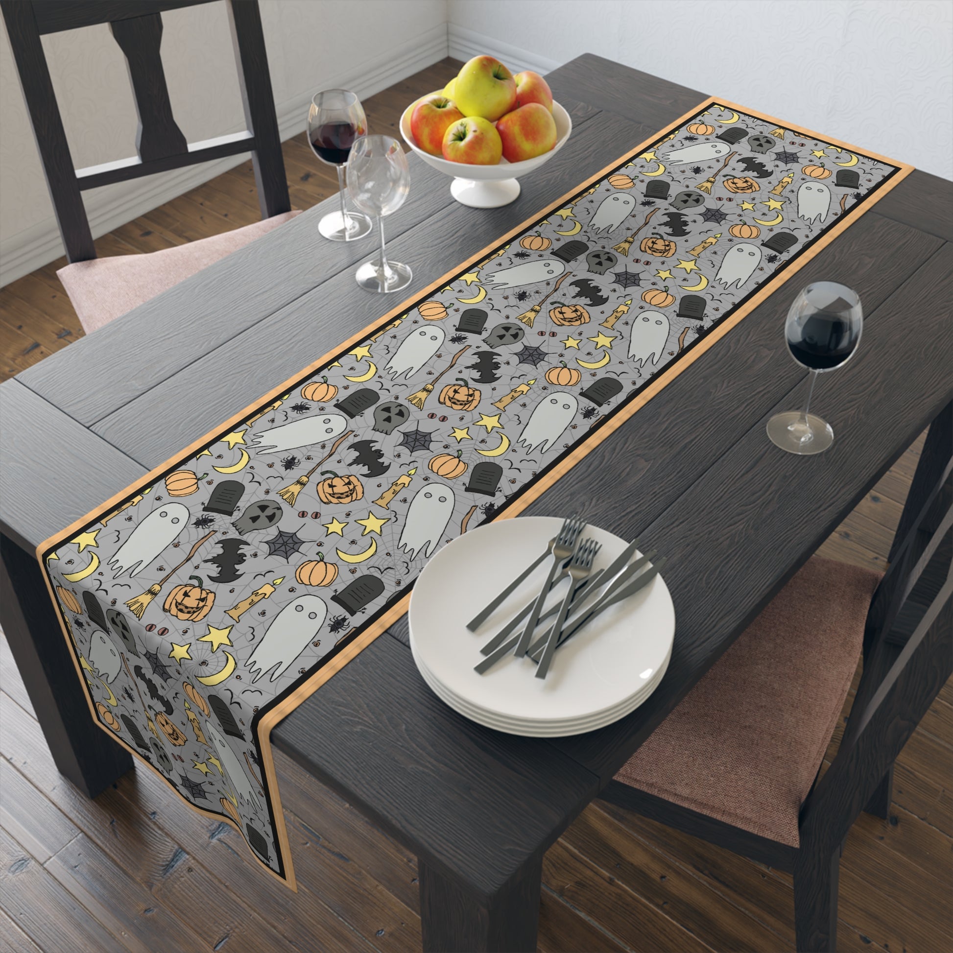 Halloween-themed table runner with ghosts, pumpkins, and bats with a gray background on front of table runner, on a wooden table.