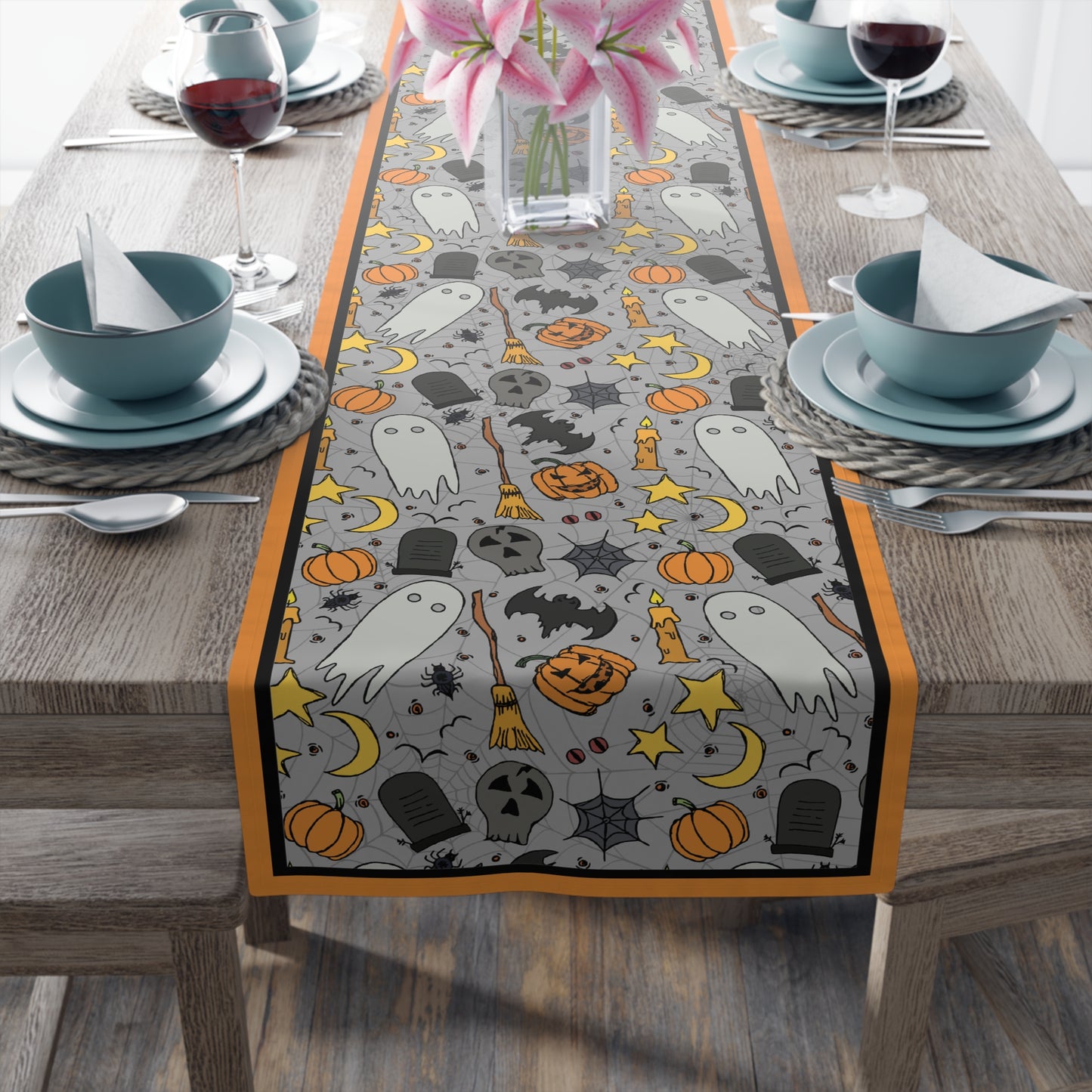Halloween-themed table runner with ghosts, pumpkins, and bats with a gray background on front of table runner, on a wooden table with place settings.