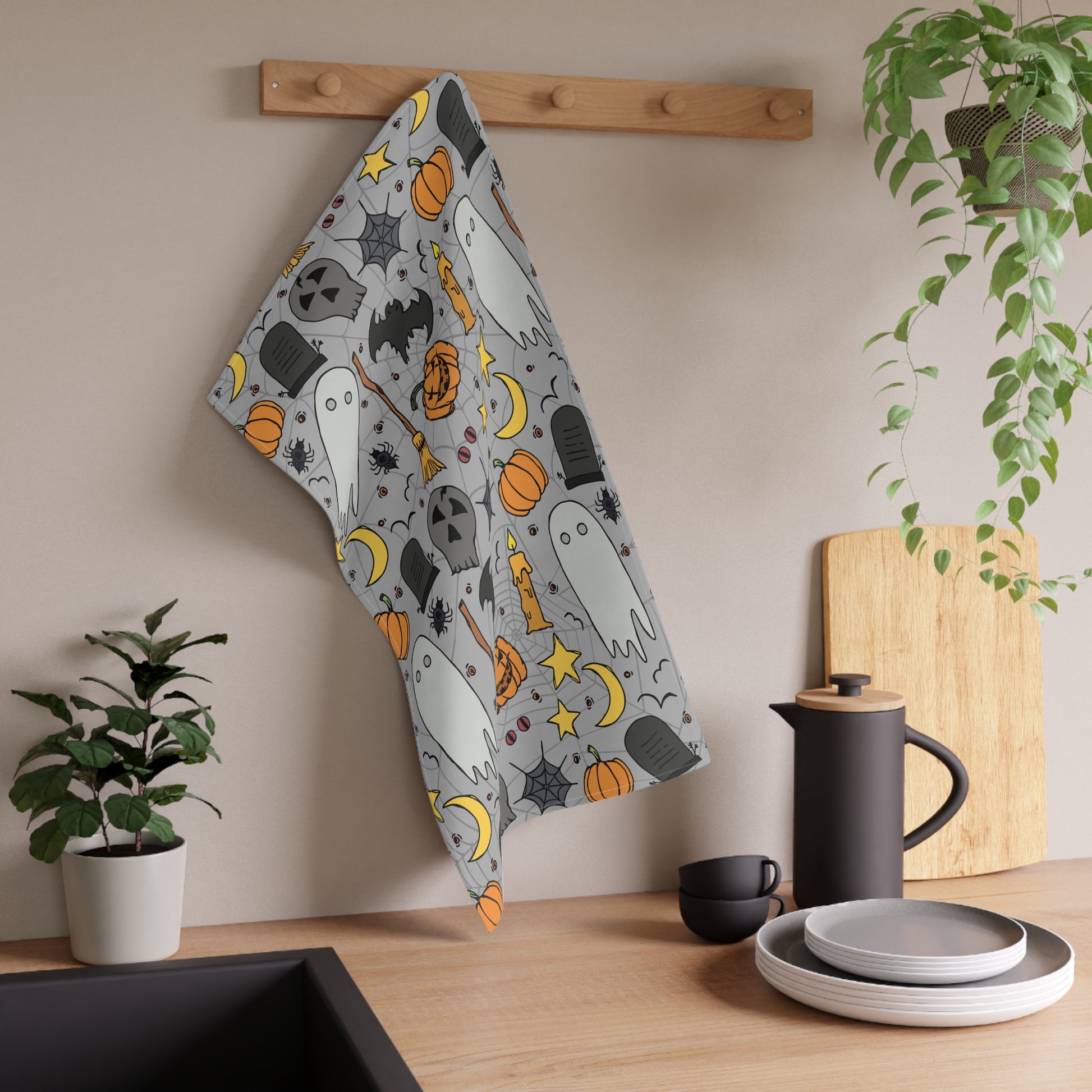 Halloween-themed tea towel with ghosts, pumpkins, and bats on a gray background printed on front of kitchen towel, hanging on a rack in a kitchen.