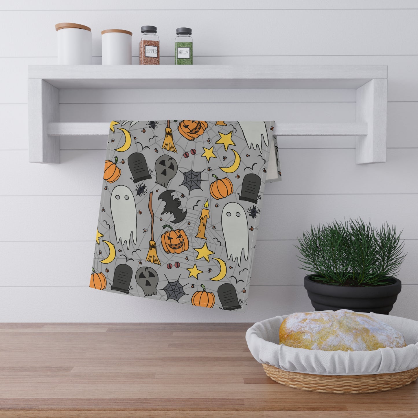 Halloween-themed tea towel with ghosts, pumpkins, and bats with a gray background printed on front of kitchen towel, hanging on a wooden shelf with decorative items.