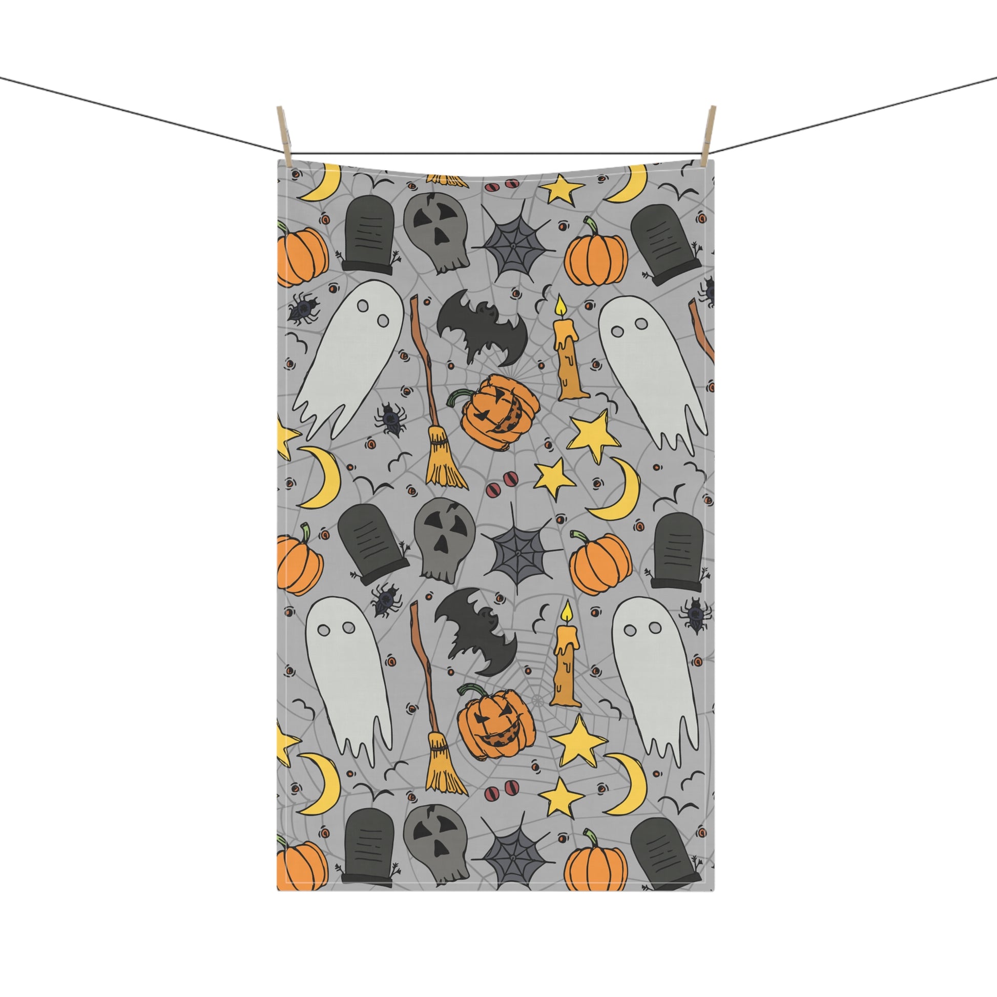 Halloween-themed tea towel with ghosts, pumpkins, and bats on a gray background printed on front of kitchen towel, hanging on a line.
