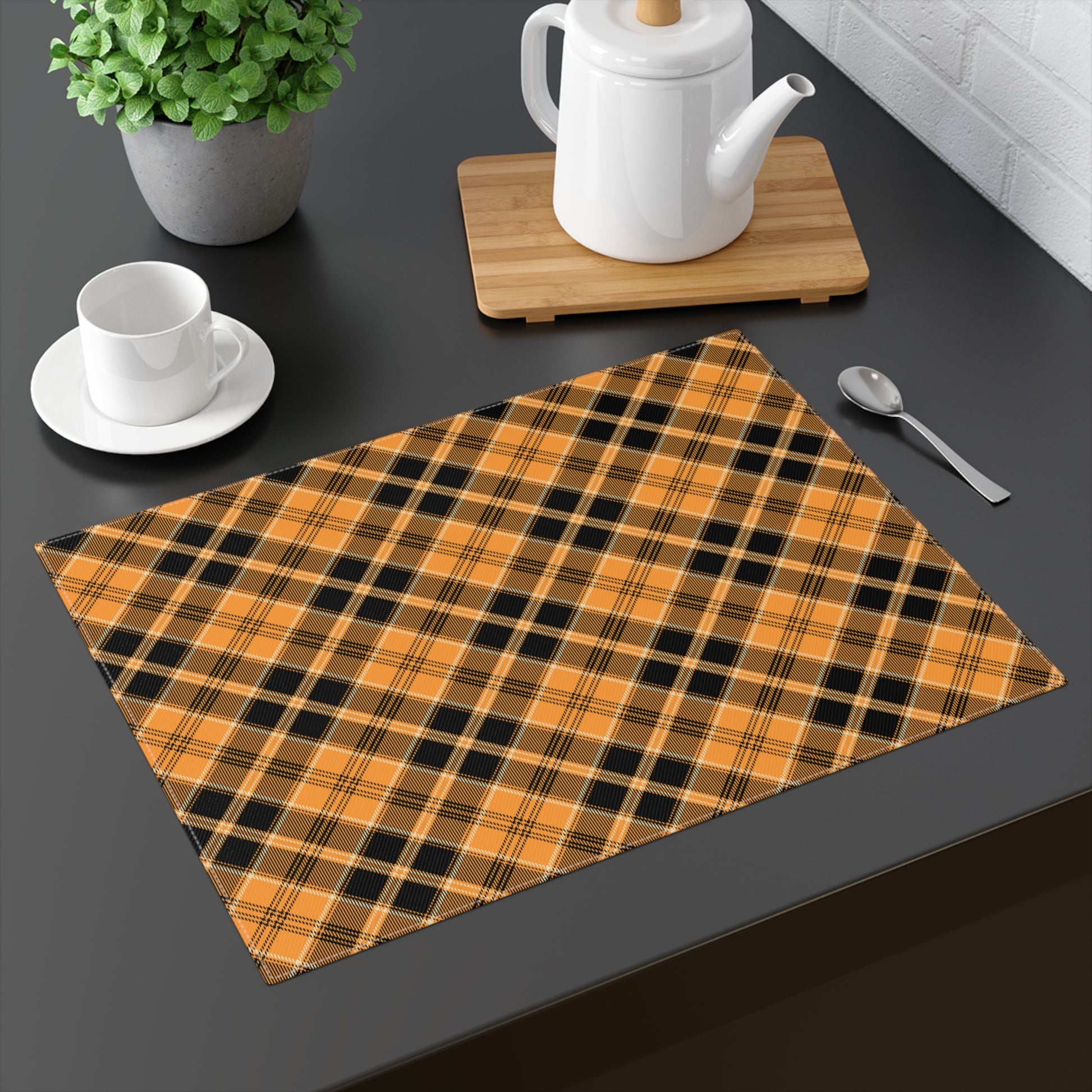 Halloween Plaid placemat has plaid pattern in orange and black colors printed on top of placemat, on a dark table with a teapot, cup, and spoon.