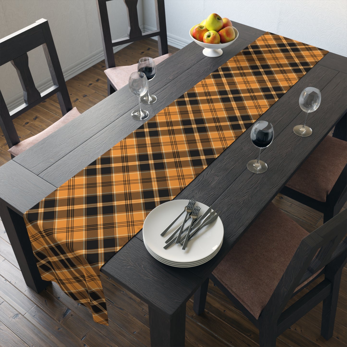 Pumpkin plaid table runner has a black and orange plaid pattern on front of table runner, on a dining table  with plates, and glasses in a room.