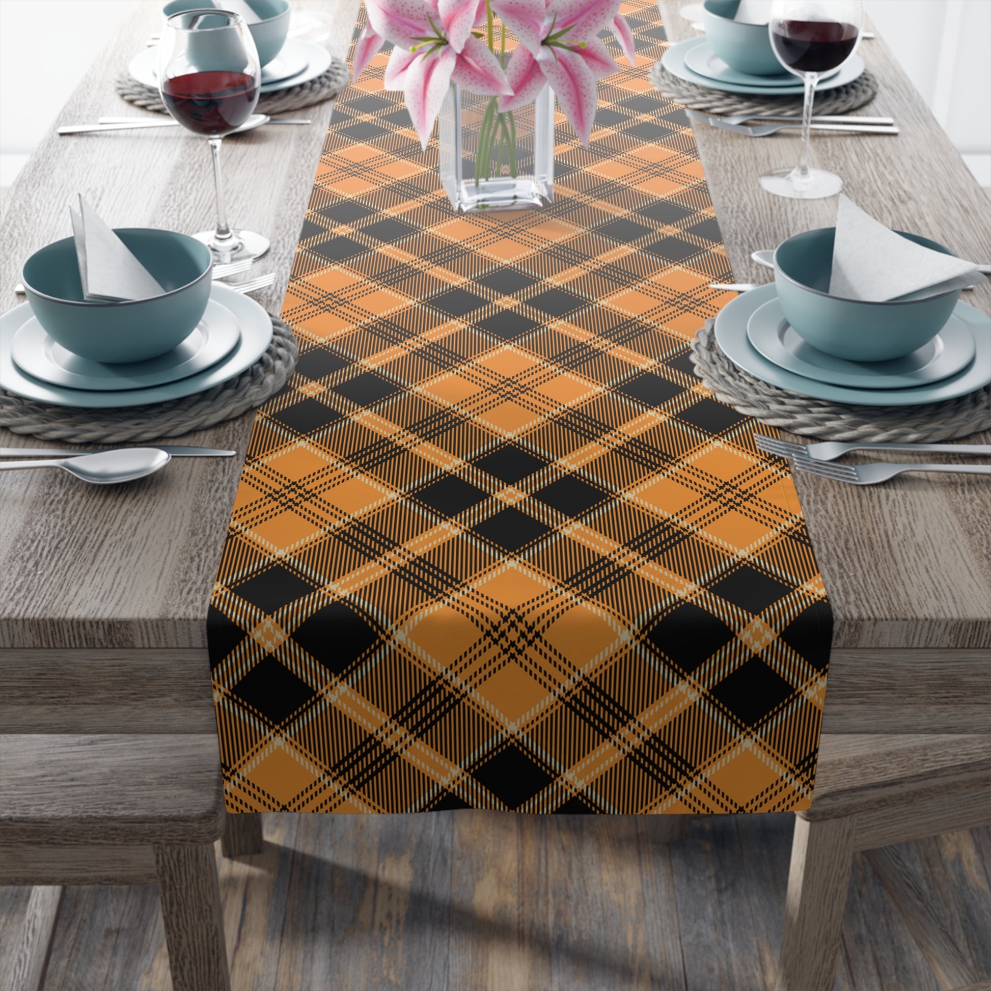 Pumpkin plaid table runner has a black and orange plaid pattern on front of table runner, on a wooden table with place settings