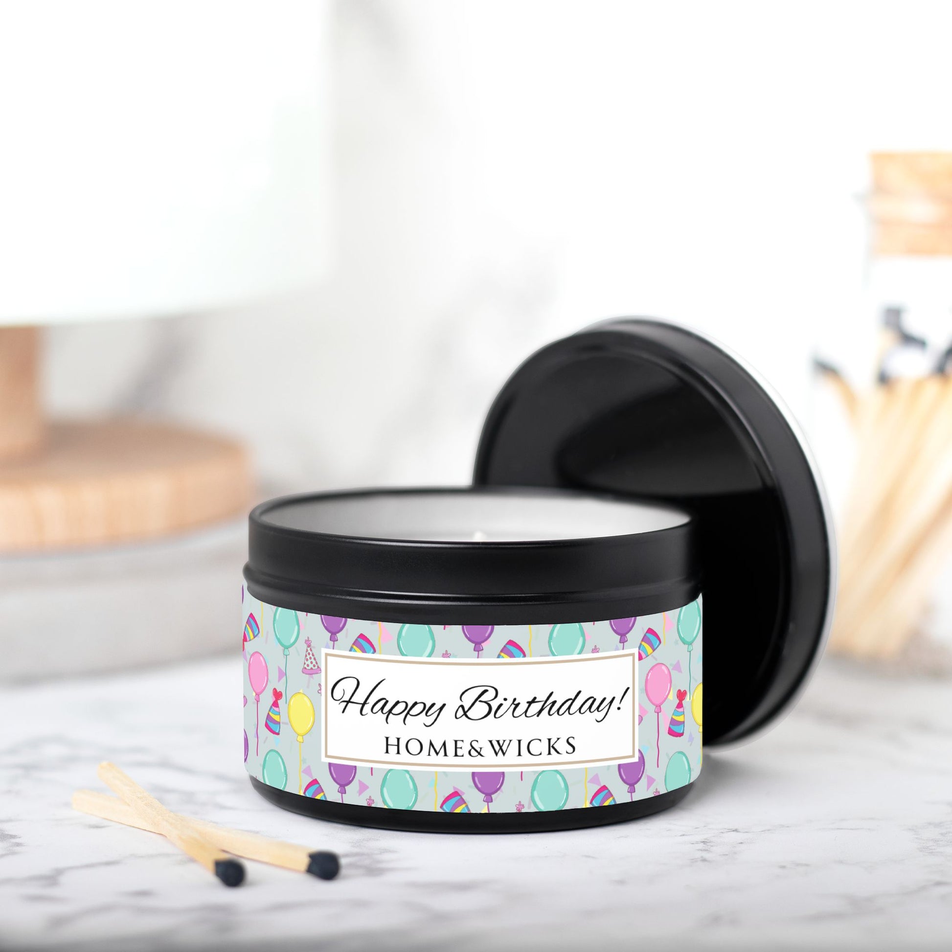 Black candle tin with 'Happy Birthday' label with colorful balloon, birthday hats and confetti design and 'Home & Wicks' branding on a light background.with match sticks