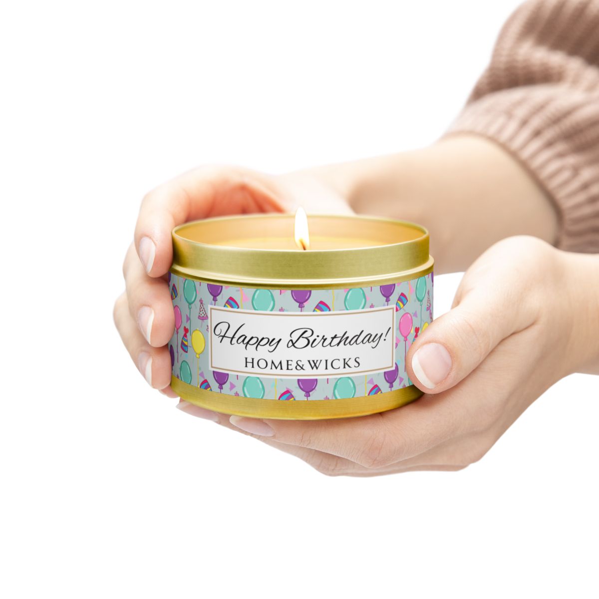 Hand holding lit gold candle tin with 'Happy Birthday' label with colorful balloon, birthday hats and confetti design on a white background
