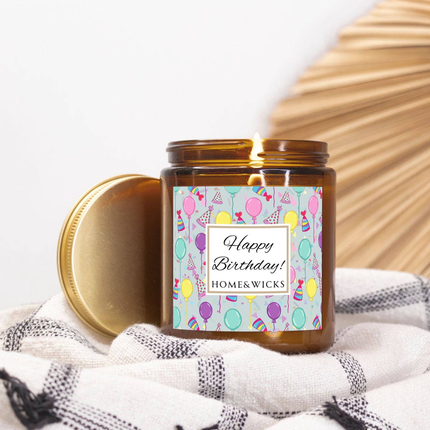 Amber candle jar with gold label with a 'Happy Birthday' label and colorful design with birthday balloons, birthday hats and confetti on a white background on a white and black patterned fabric background.