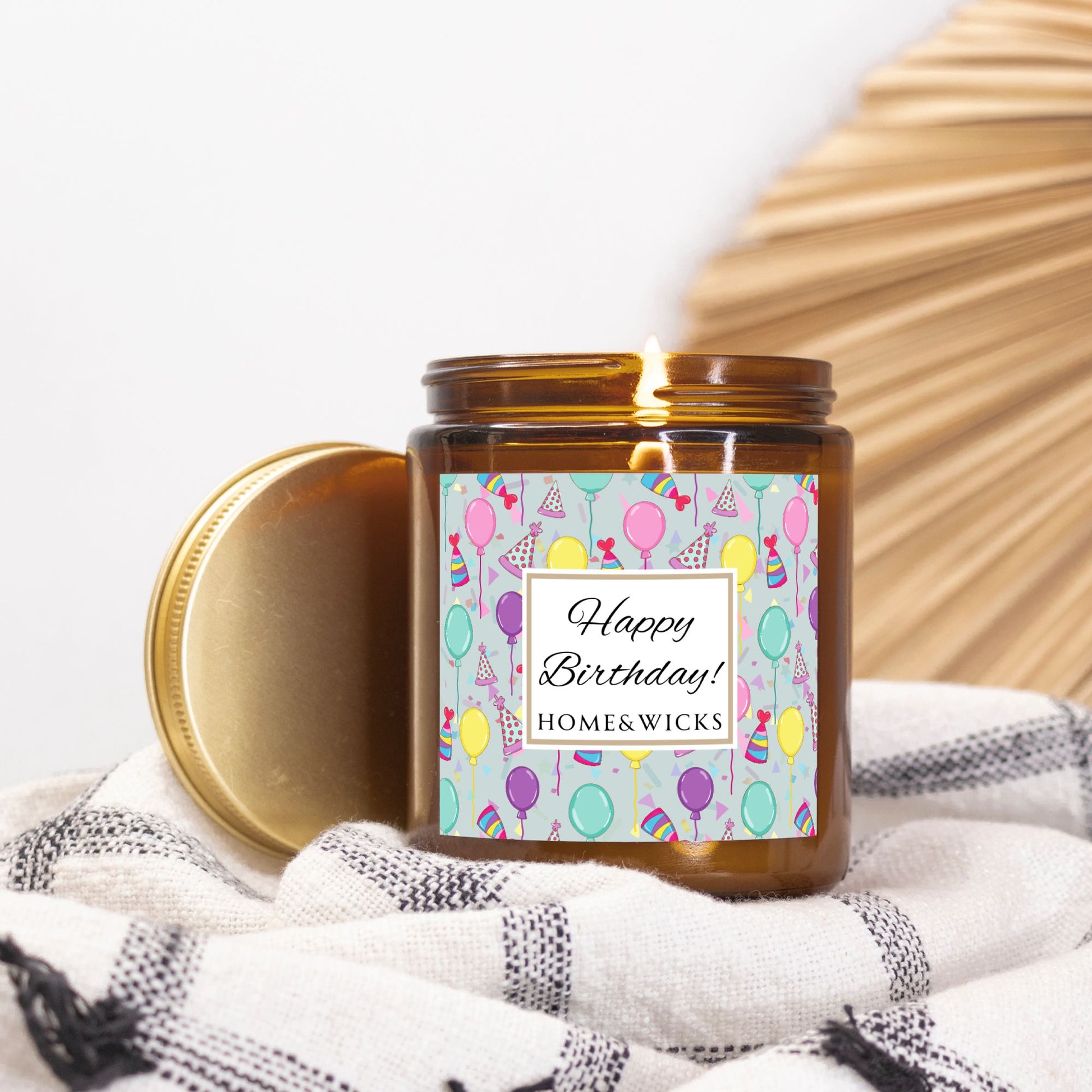 Amber candle jar with gold label with a 'Happy Birthday' label and colorful design with birthday balloons, birthday hats and confetti on a white background on a white and black patterned fabric background.