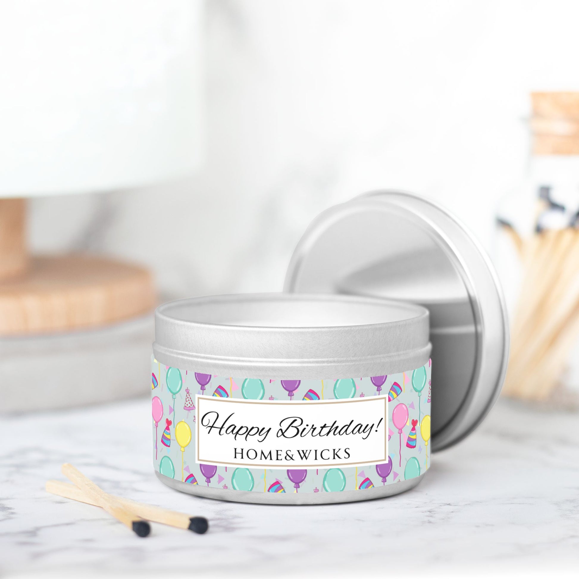 Silver candle tin with 'Happy Birthday' label and colorful balloon, birthday hats and confetti design label and matchbox on a marble surface