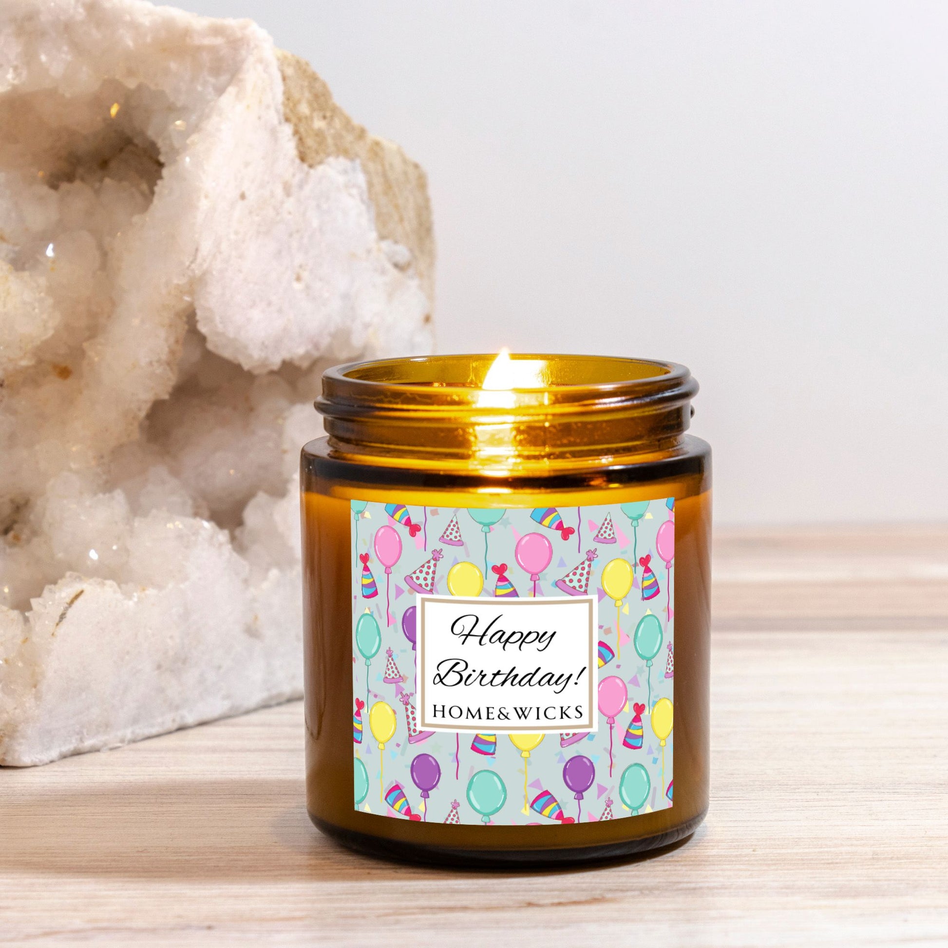 Amber candle jar with gold lid with a 'Happy Birthday' label featuring balloons, birthday party hats and confetti on a wooden surface with a white background