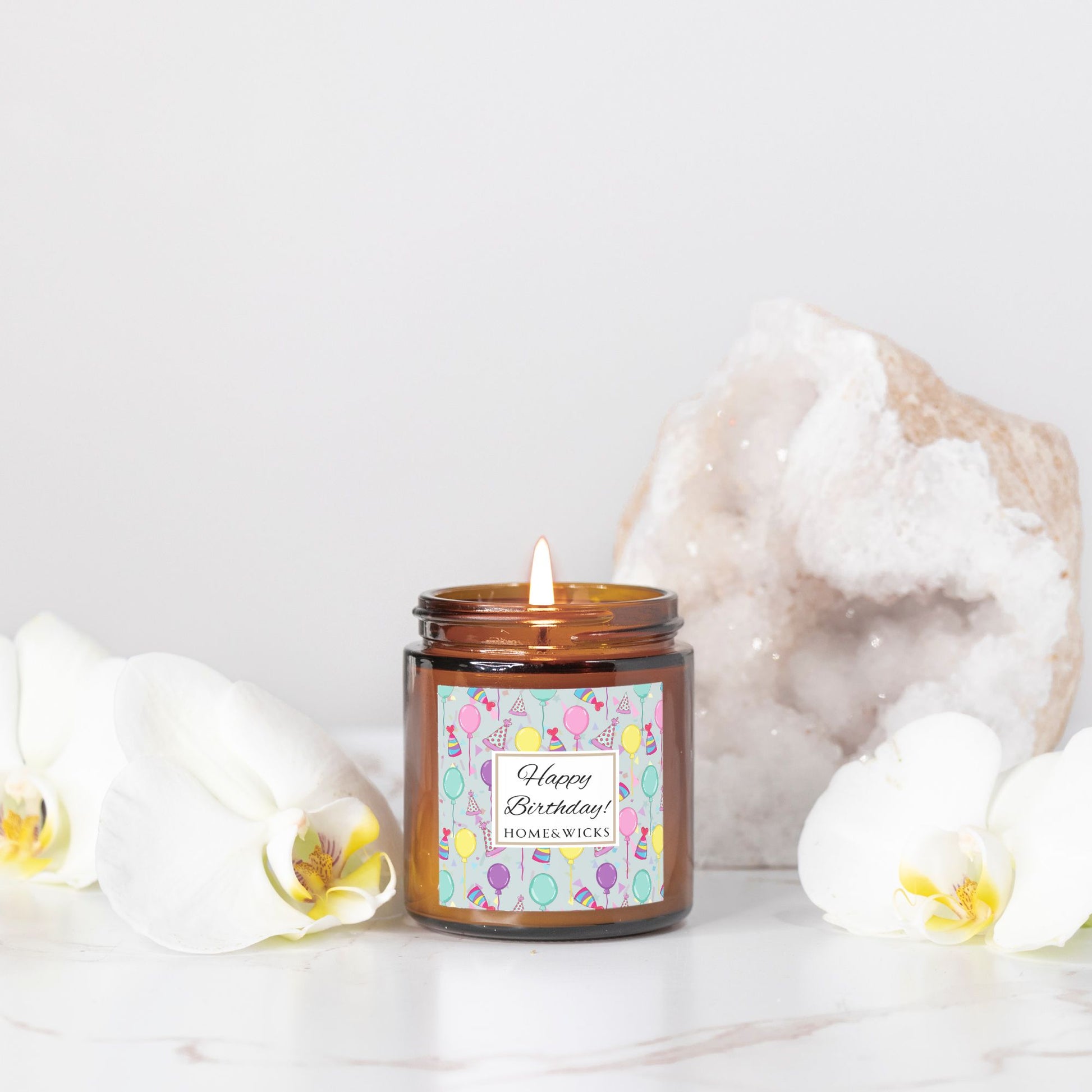 Amber candle jar with gold lid with a 'Happy Birthday' label featuring balloons, birthday party hats and confetti on a light background.