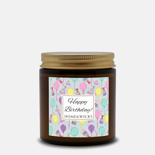 Amber candle jar with gold lid with a 'Happy Birthday' label featuring balloons, birthday party hats and confetti on a white background.
