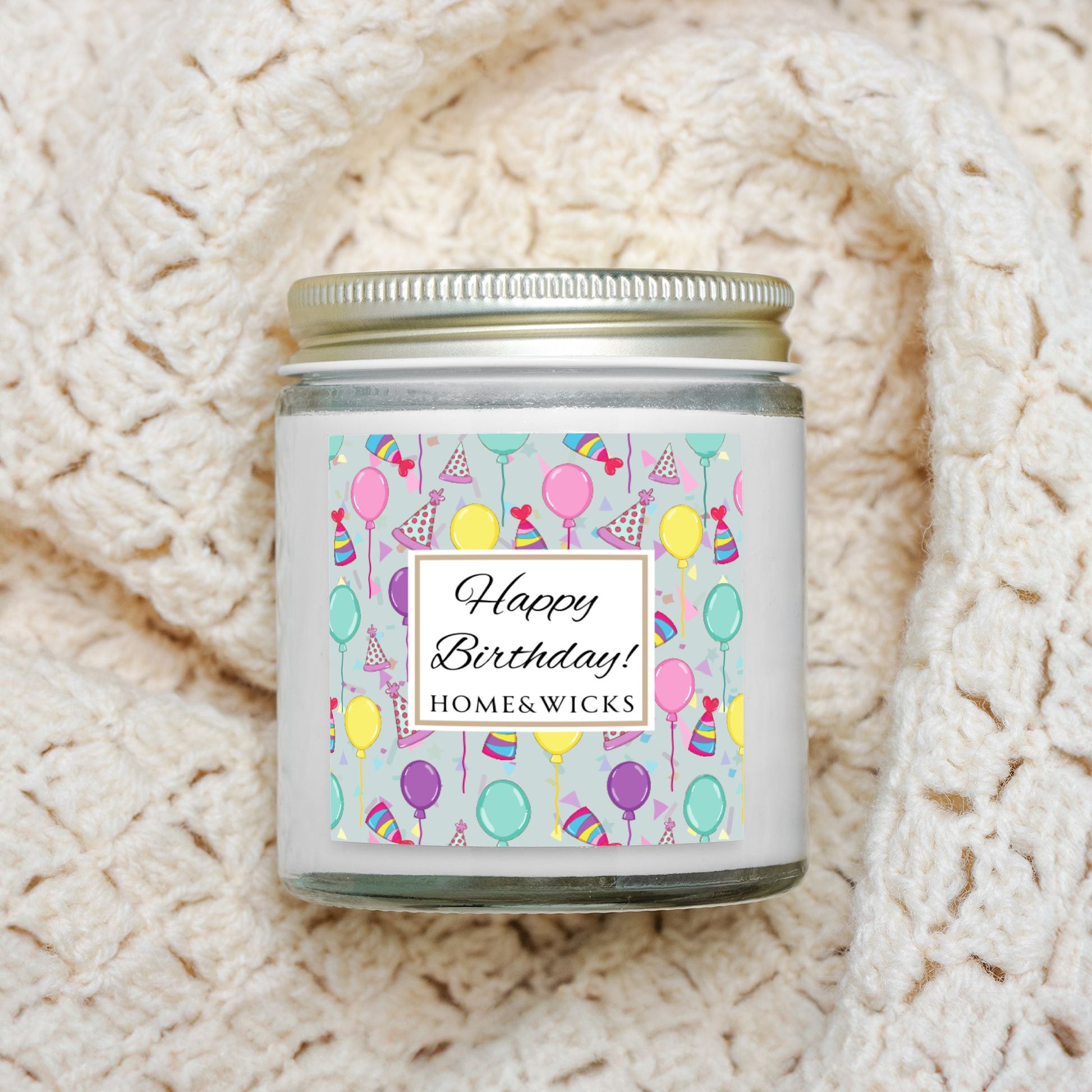 Clear candle jar with gold lid with a 'Happy Birthday' label featuring balloons, birthday party hats and confetti on a textured white background