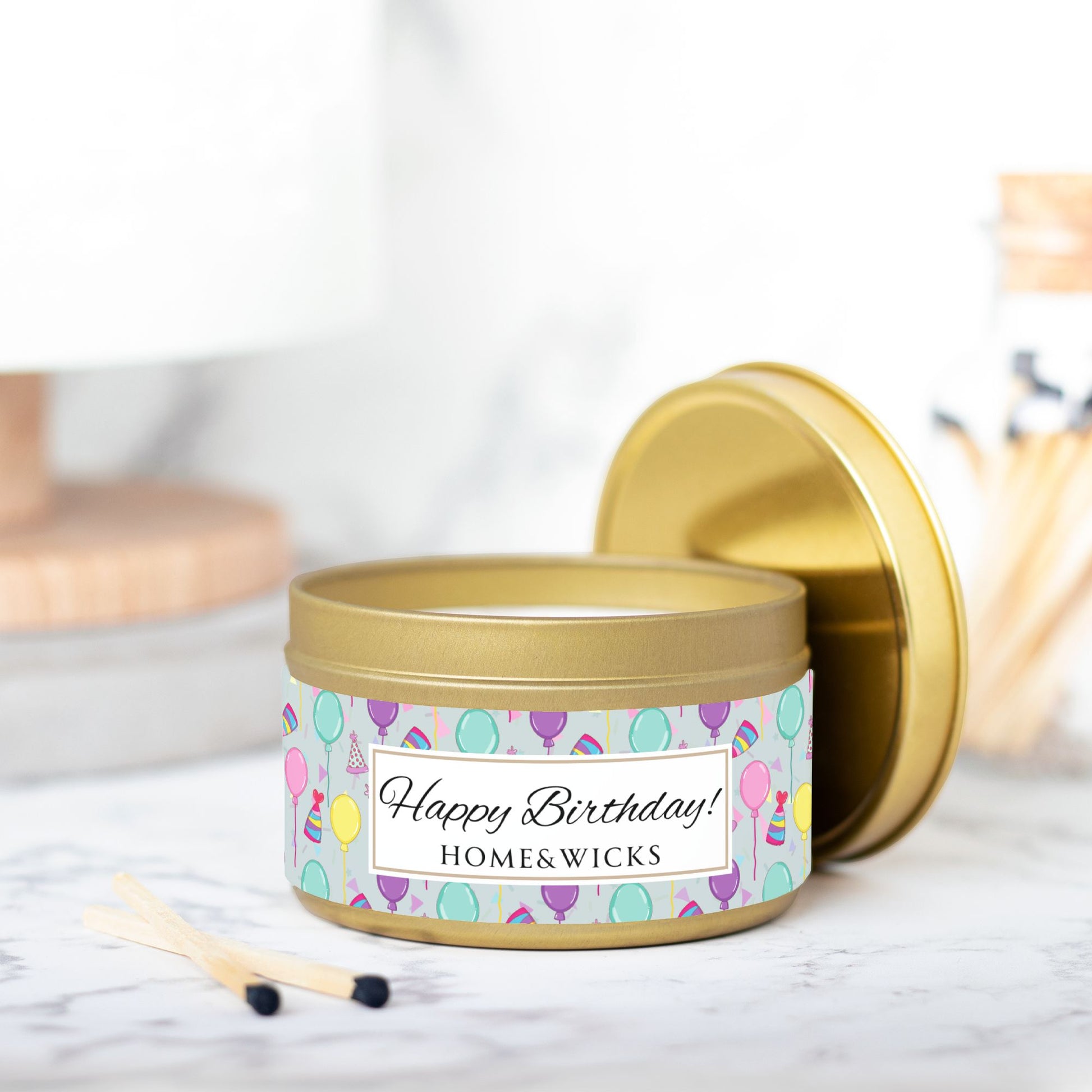 Gold candle tin with 'Happy Birthday' label with colorful balloon, birthday hats and confetti design, on a light background with match sticks