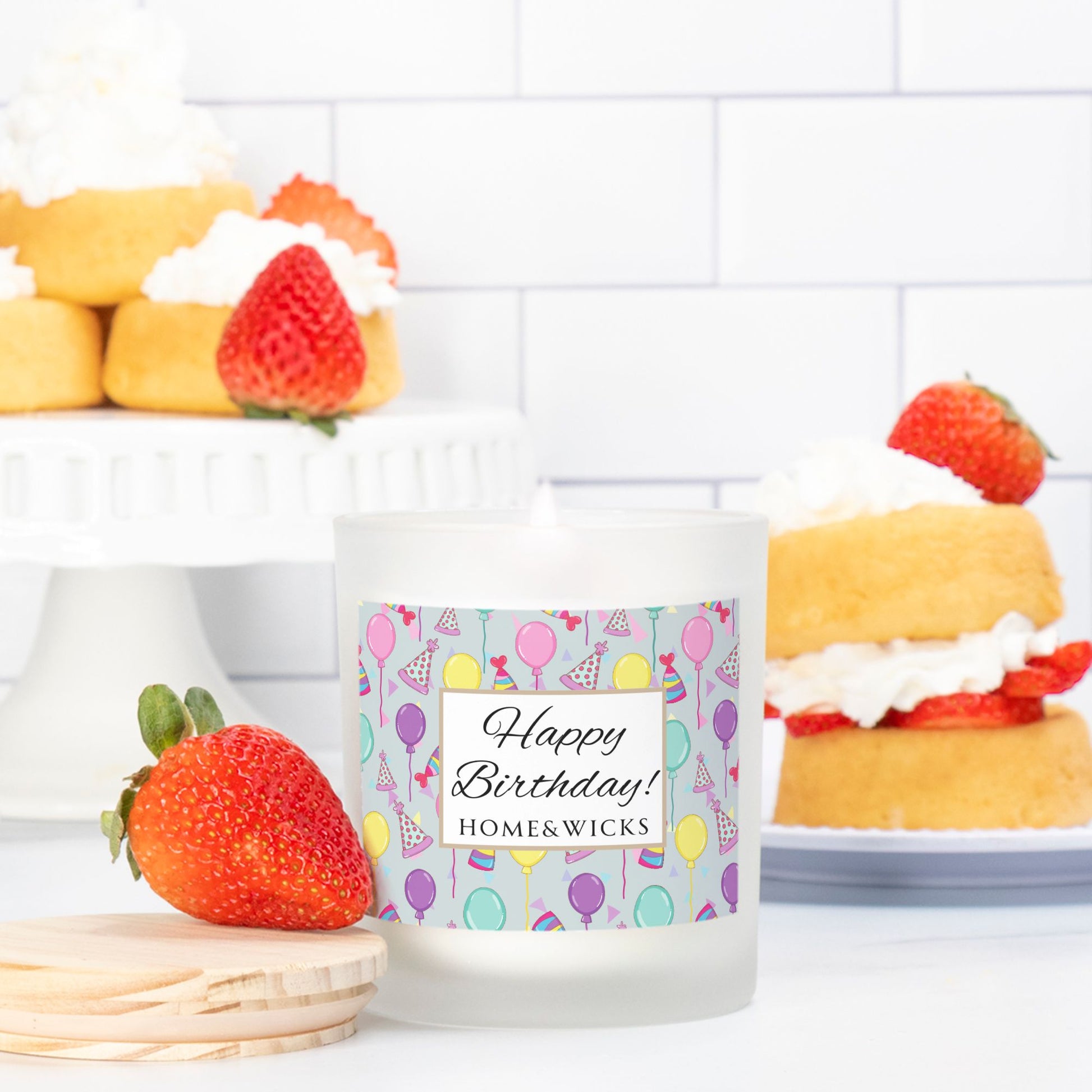 Pink wick candle in a frosted jar with bamboo wood lid and with a 'Happy Birthday' label with birthday balloons, birthday hats and confetti by Home & Wicks on a white background surrounded by strawberry shortcakes on a white tiled background