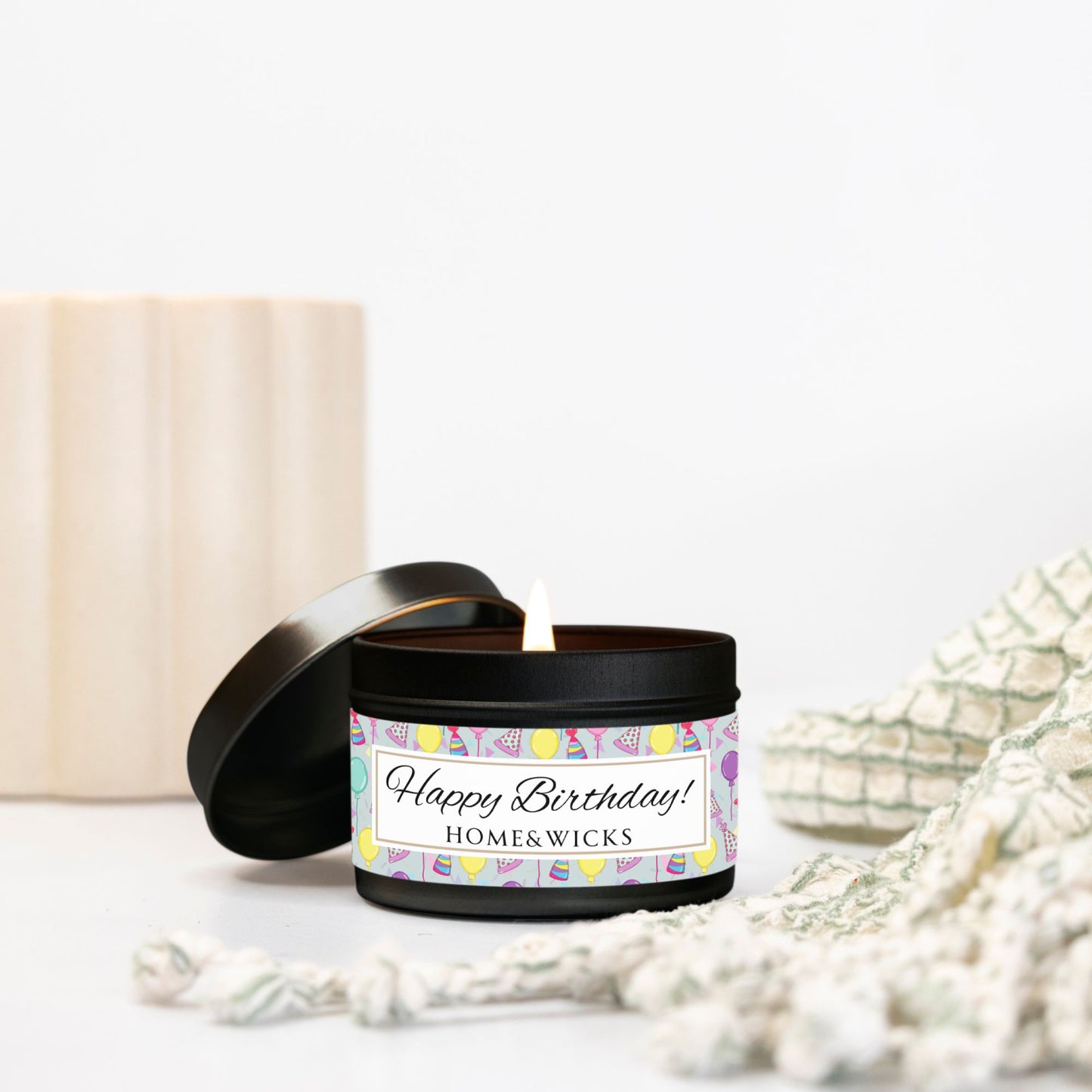 Opened cotton wicked black 4 oz. candle tin with 'Happy Birthday' label with colorful design of birthday hats, confetti and balloons placed on a white surface with decorative elements.
