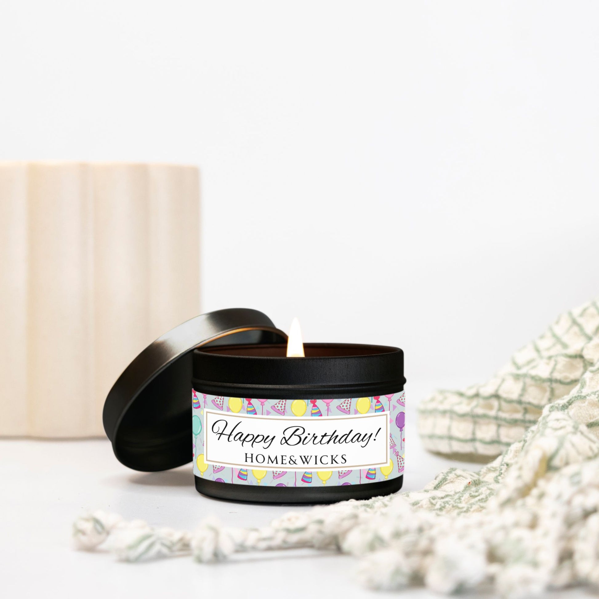 Opened cotton wicked black 4 oz. candle tin with 'Happy Birthday' label with colorful design of birthday hats, confetti and balloons placed on a white surface with decorative elements.