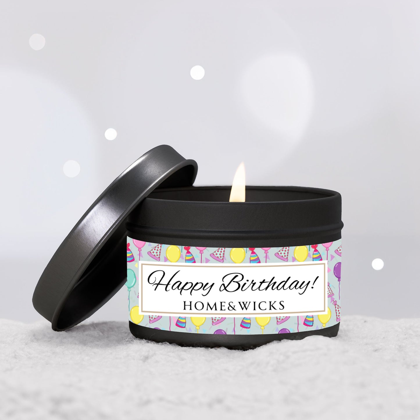 Opened cotton wicked black 4 oz. candle tin with 'Happy Birthday' label with colorful design of birthday hats, confetti and balloons by HOME&WICKS on a light gray background