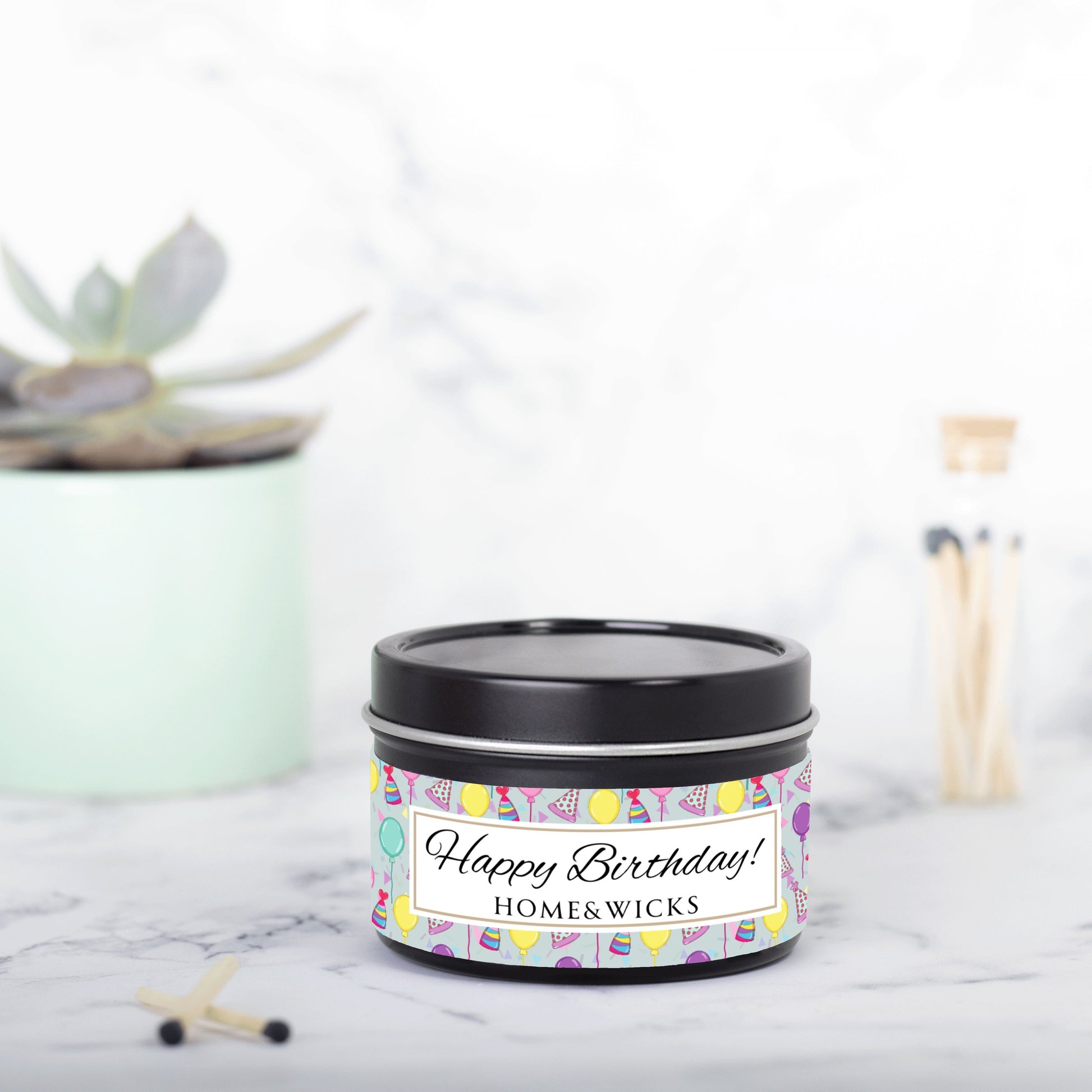 Opened cotton wicked black 4 oz. candle tin with 'Happy Birthday' label with colorful design of birthday hats, confetti and balloons by Home & wicks on a marble surface.