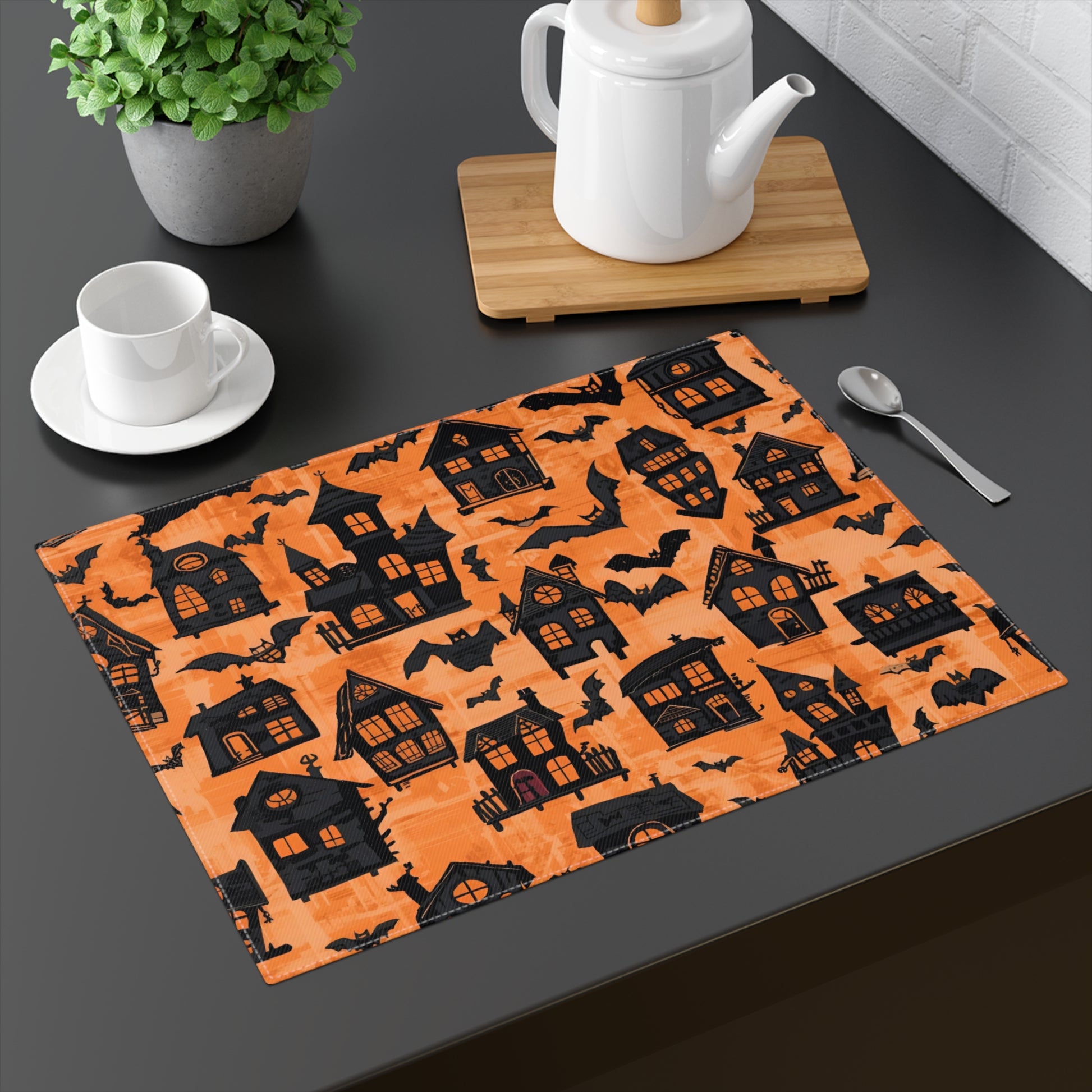 Halloween-themed placemat with black haunted houses and bats on an orange background. The placemat is placed on a dark surface with a teapot and cup.
