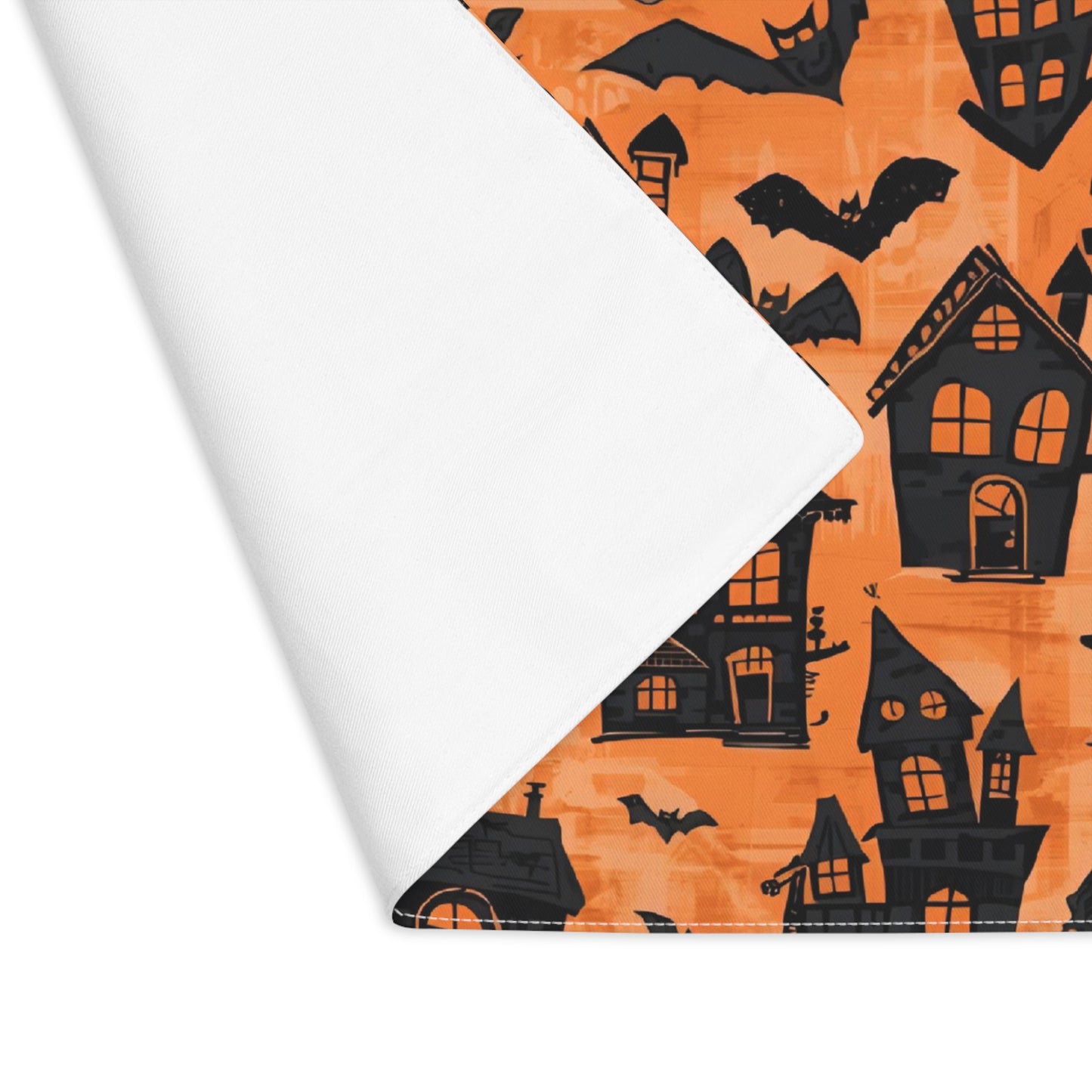 Halloween-themed fabric placemat with black haunted houses and bats on orange background. The placemat's corner is folded over to reveal white backside.