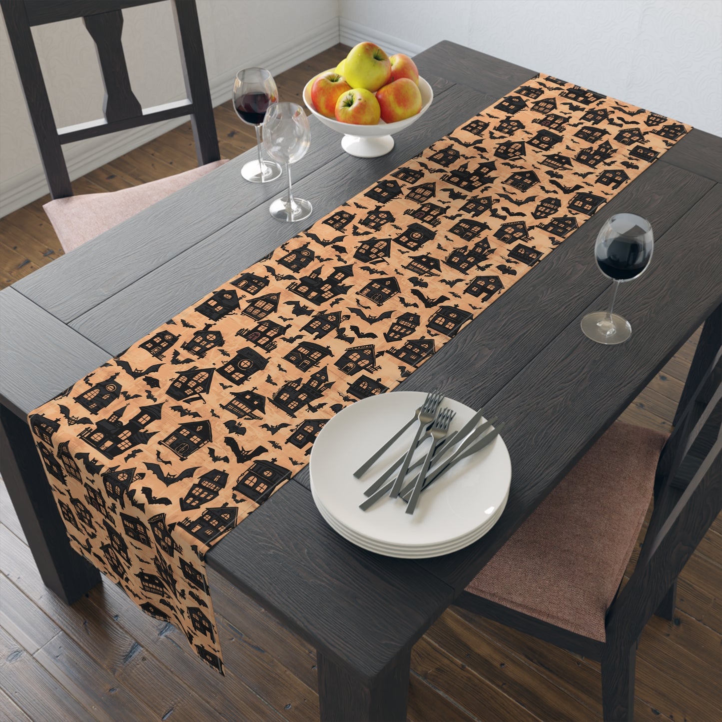 Black Haunted Houses table runner with black haunted houses and bats printed on an orange background design on the table runner sits on a dining table with plates, and cutlery.