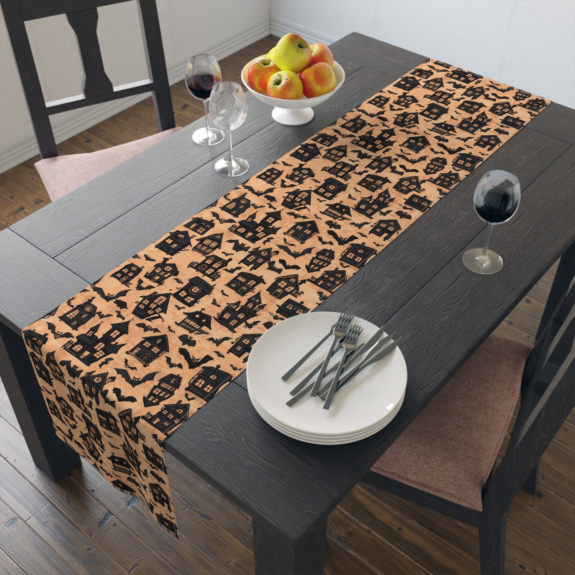 Black Haunted Houses table runner with black haunted houses and bats printed on an orange background design on the table runner sits on a dining table with plates, and cutlery.