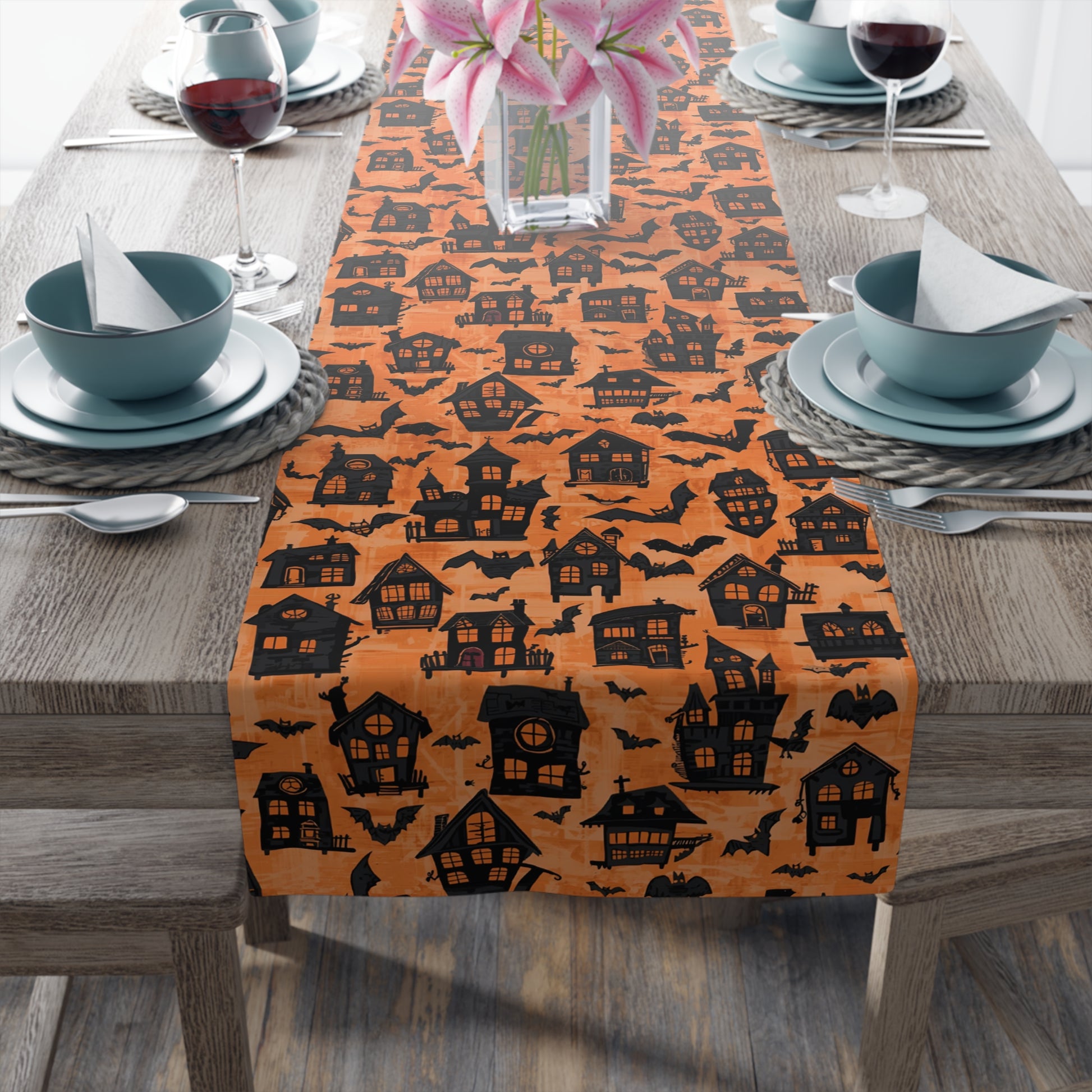 Halloween-themed table runner with black haunted houses pattern on an orange background, placed on a wooden table with dishes and vase sits on this Haunted Houses table runner.