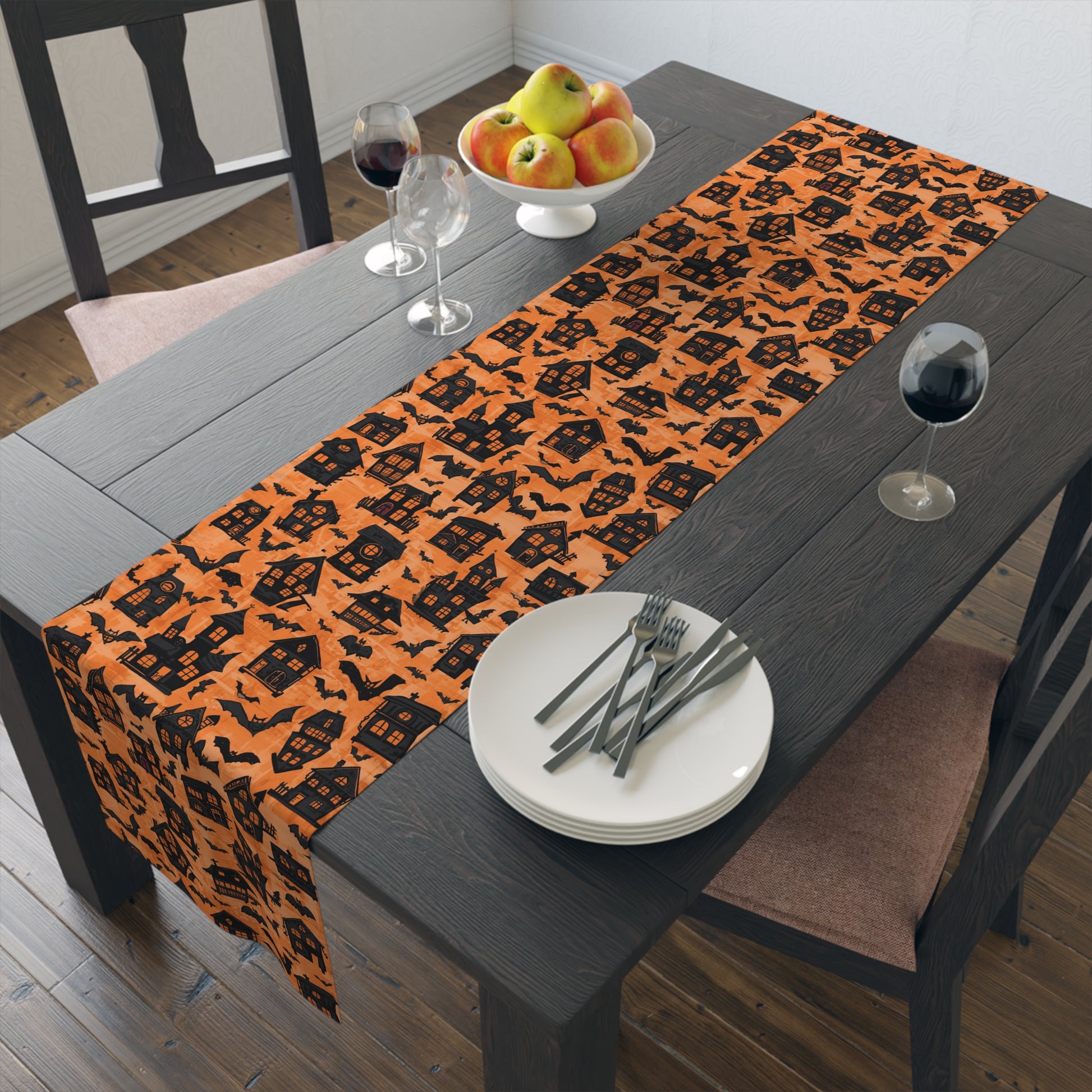 Black Haunted Houses table runner with black haunted houses and bats on an orange background design printed on the table runner that sits on a dining table with plates, and glasses.