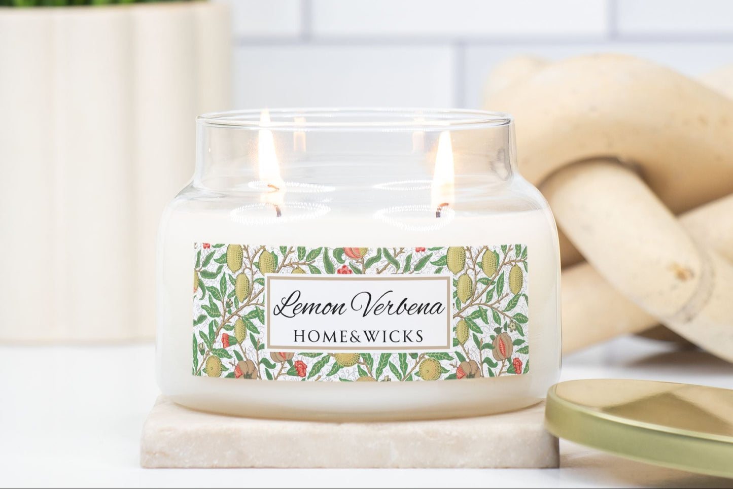 Lemon Verbena candle with decorative label on a white surface with a plant and lemon tree branches in the background