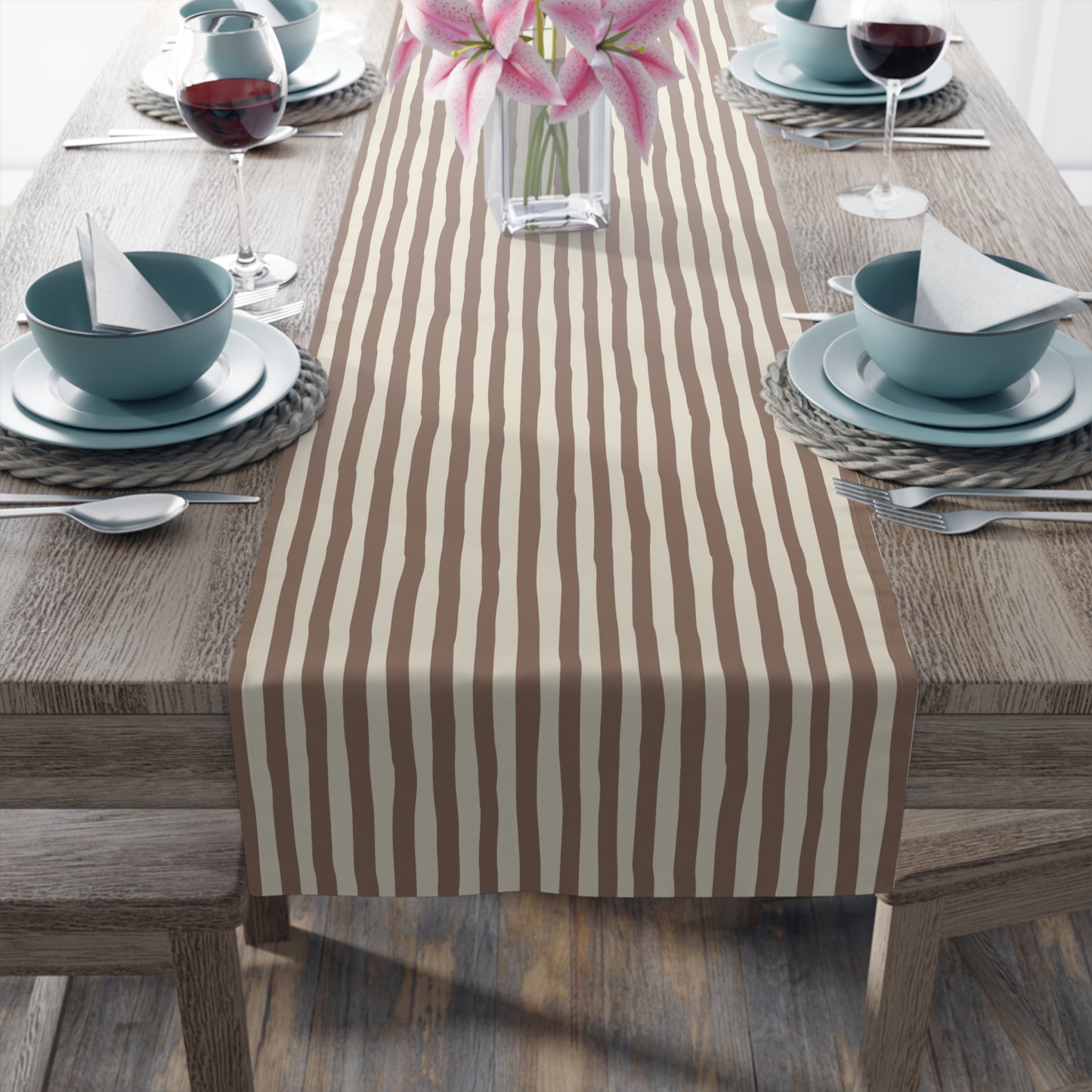 Dining table set with a brown and beige striped table runner, plates, bowls, and glasses on a wooden floor.