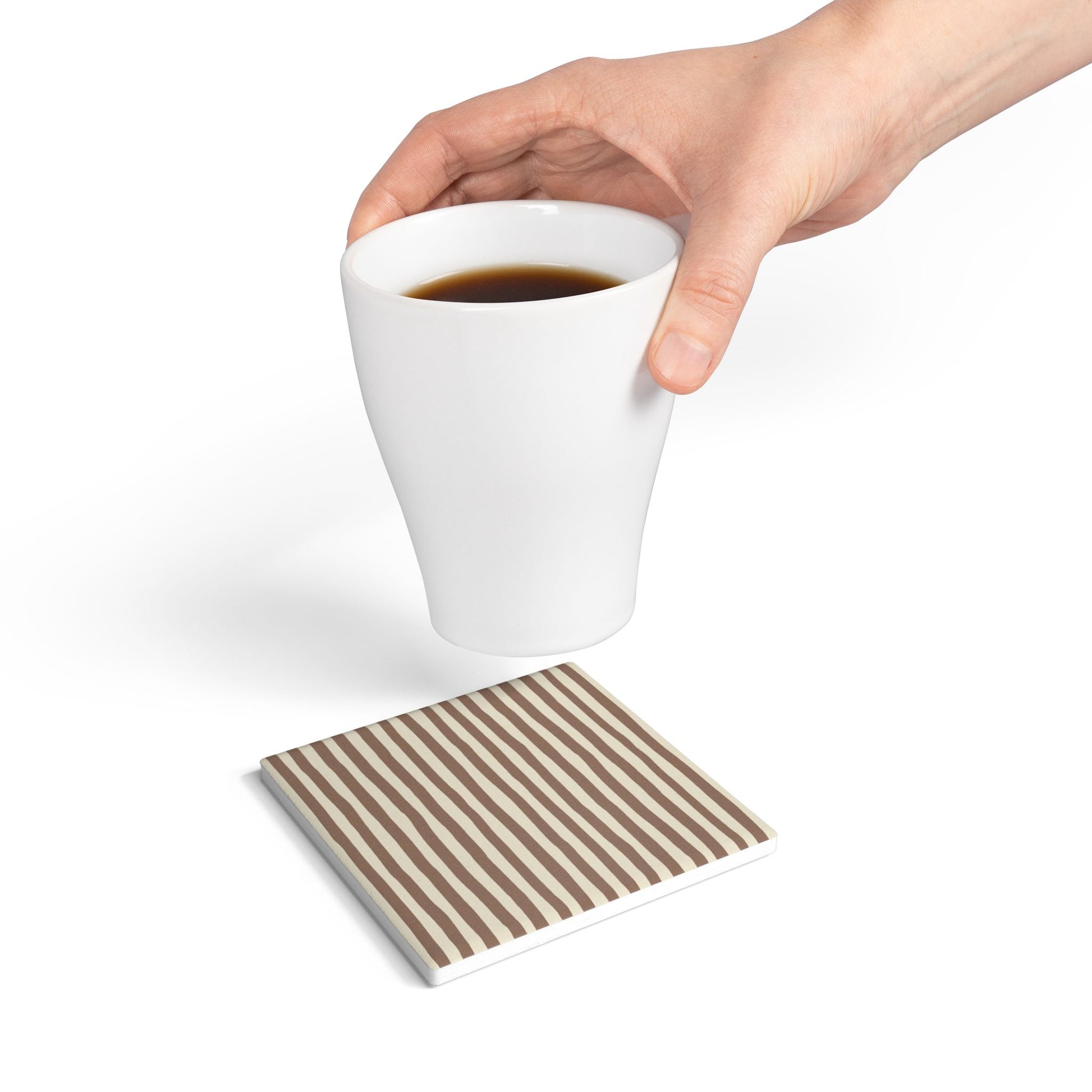 Hand holding a white mug with coffee on a mocha brown striped ceramic tile coaster.