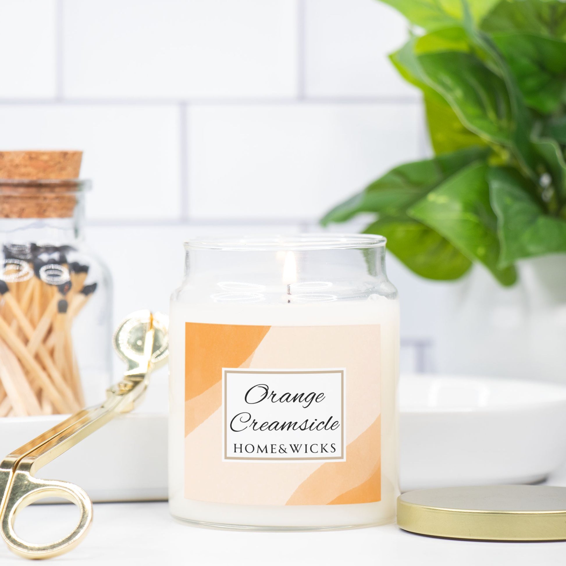 Candle jar labeled 'Orange Creamsicle' by HOME&WICKS on a white surface with a plant and matches in the background.