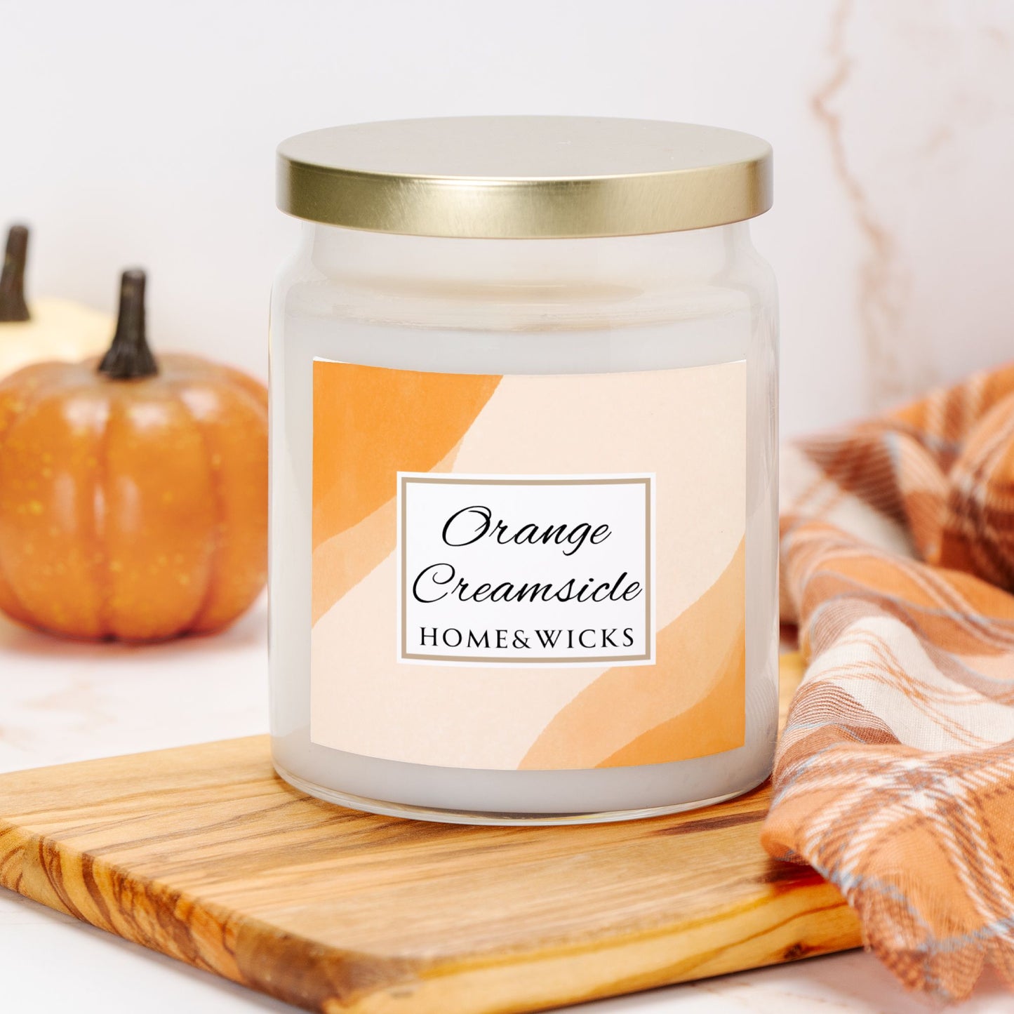 Candle jar labeled 'Orange Creamsicle' by HOME&WICKS on a wooden board with pumpkins and a plaid cloth in the background.